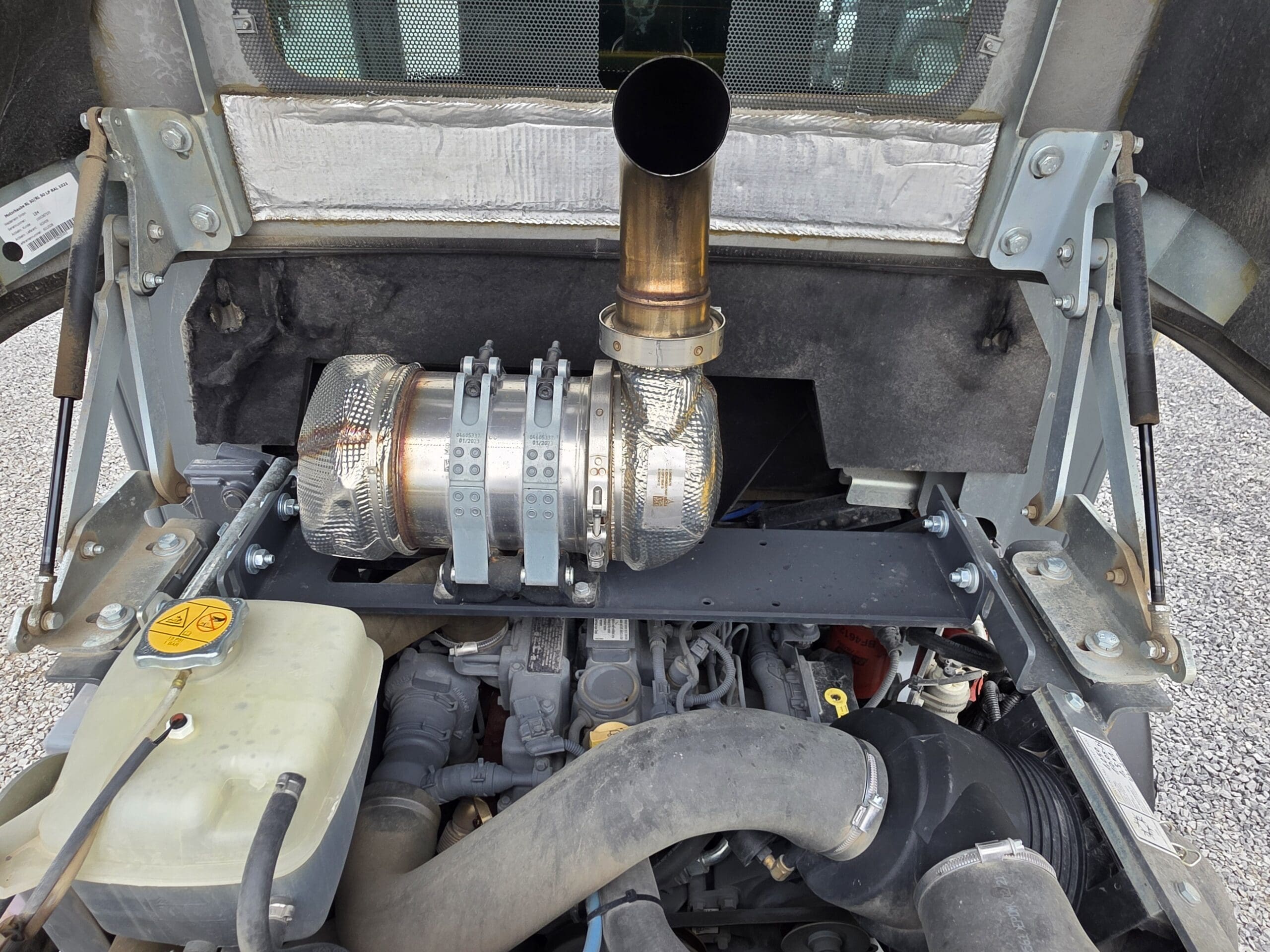 Gros plan du compartiment moteur d'un véhicule avec des composants d'échappement, des tuyaux et des pièces métalliques visibles. L'accent est mis sur un turbocompresseur ou une pièce d'échappement en métal brillant près du centre et sur un bouchon de liquide jaune à gauche.