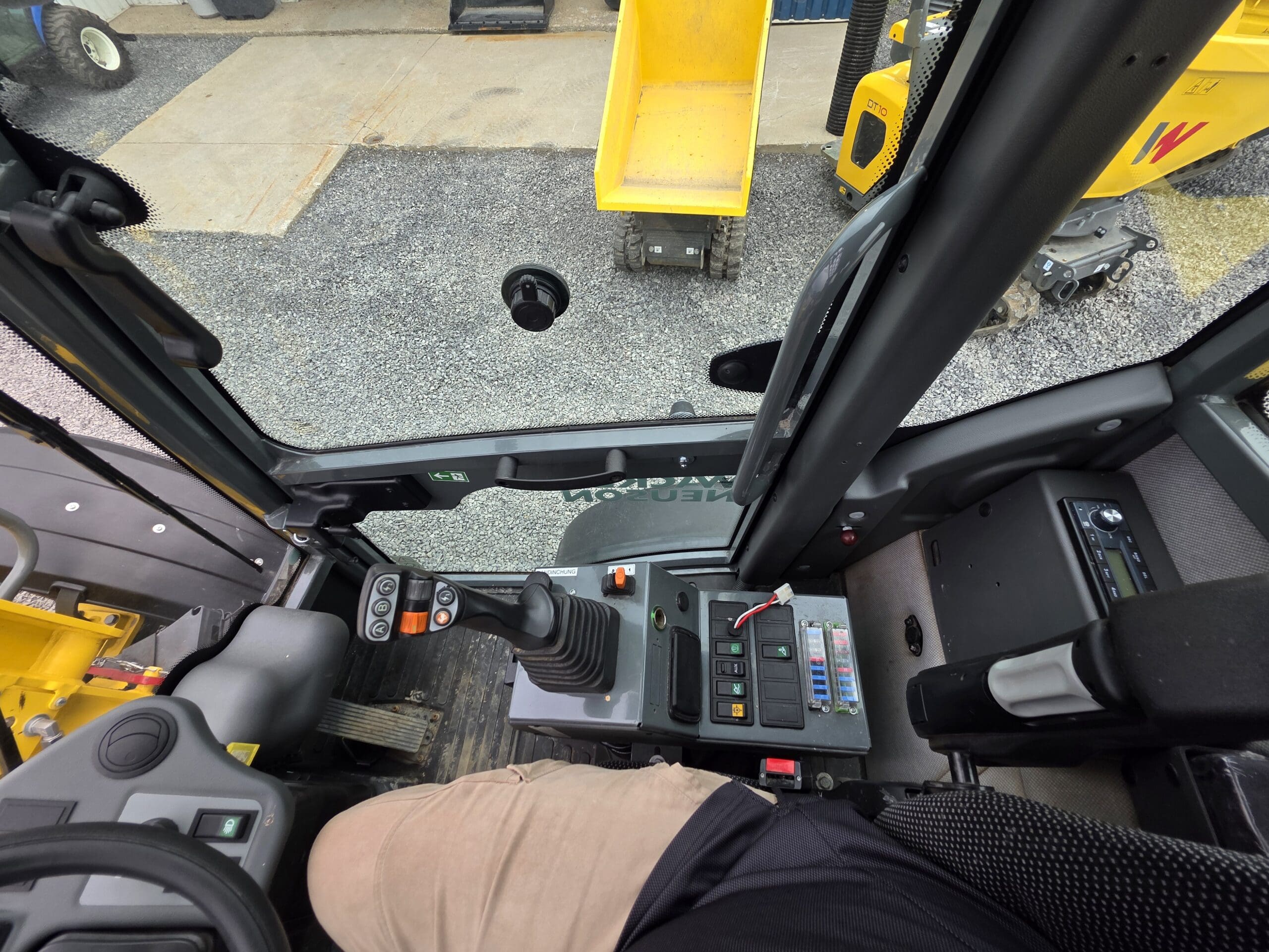 Vue de l'intérieur de la cabine d'un véhicule de construction, montrant la jambe du conducteur, le panneau de commande avec boutons et leviers, le volant et un camion-benne jaune à l'extérieur de la fenêtre.