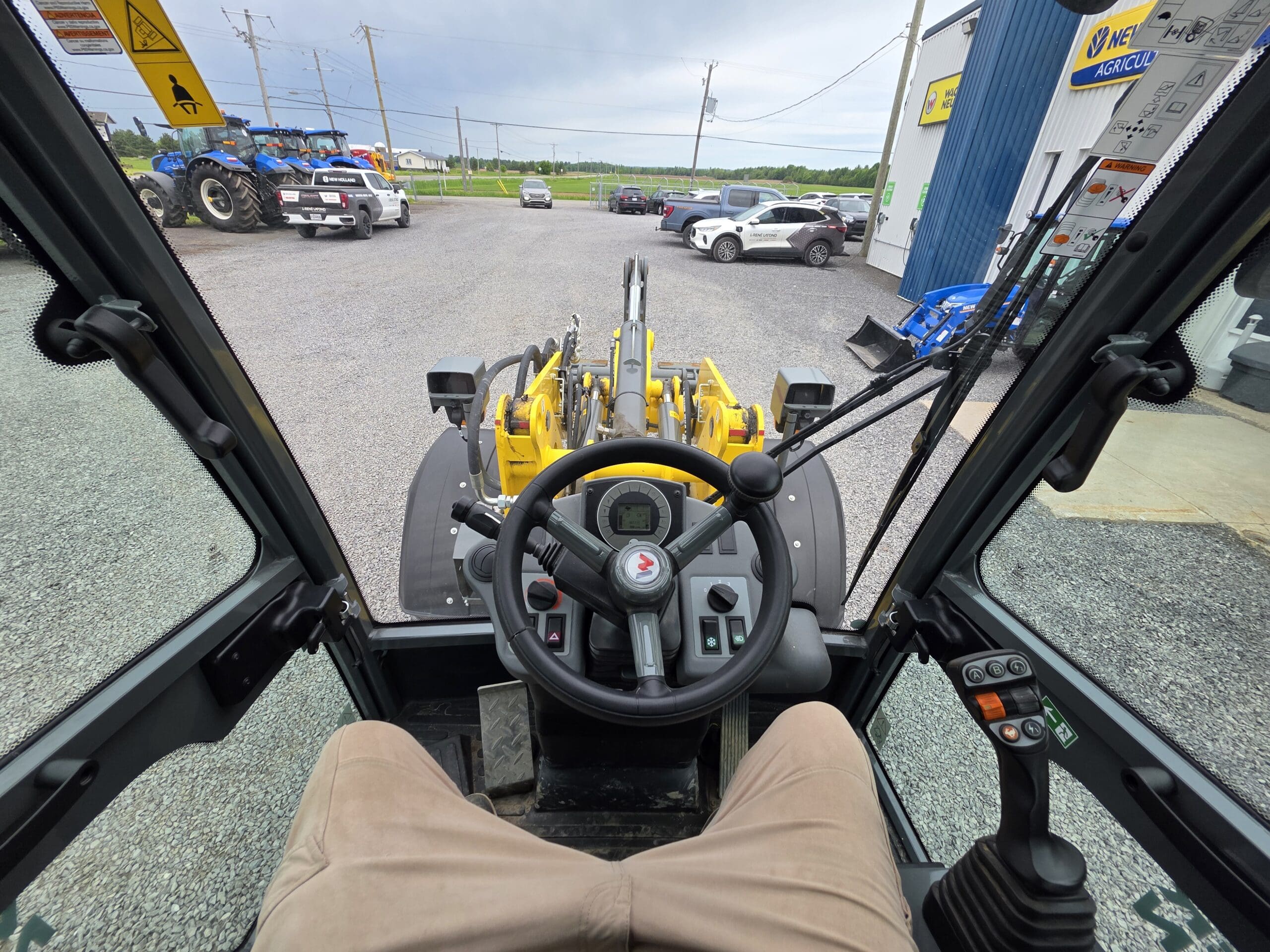 Vue de l'intérieur de la cabine d'un engin de chantier jaune, montrant le volant, les commandes et un bras de chargement à l'extérieur. Des voitures et des tracteurs sont garés sur un parking en gravier près d'un bâtiment bleu et blanc.