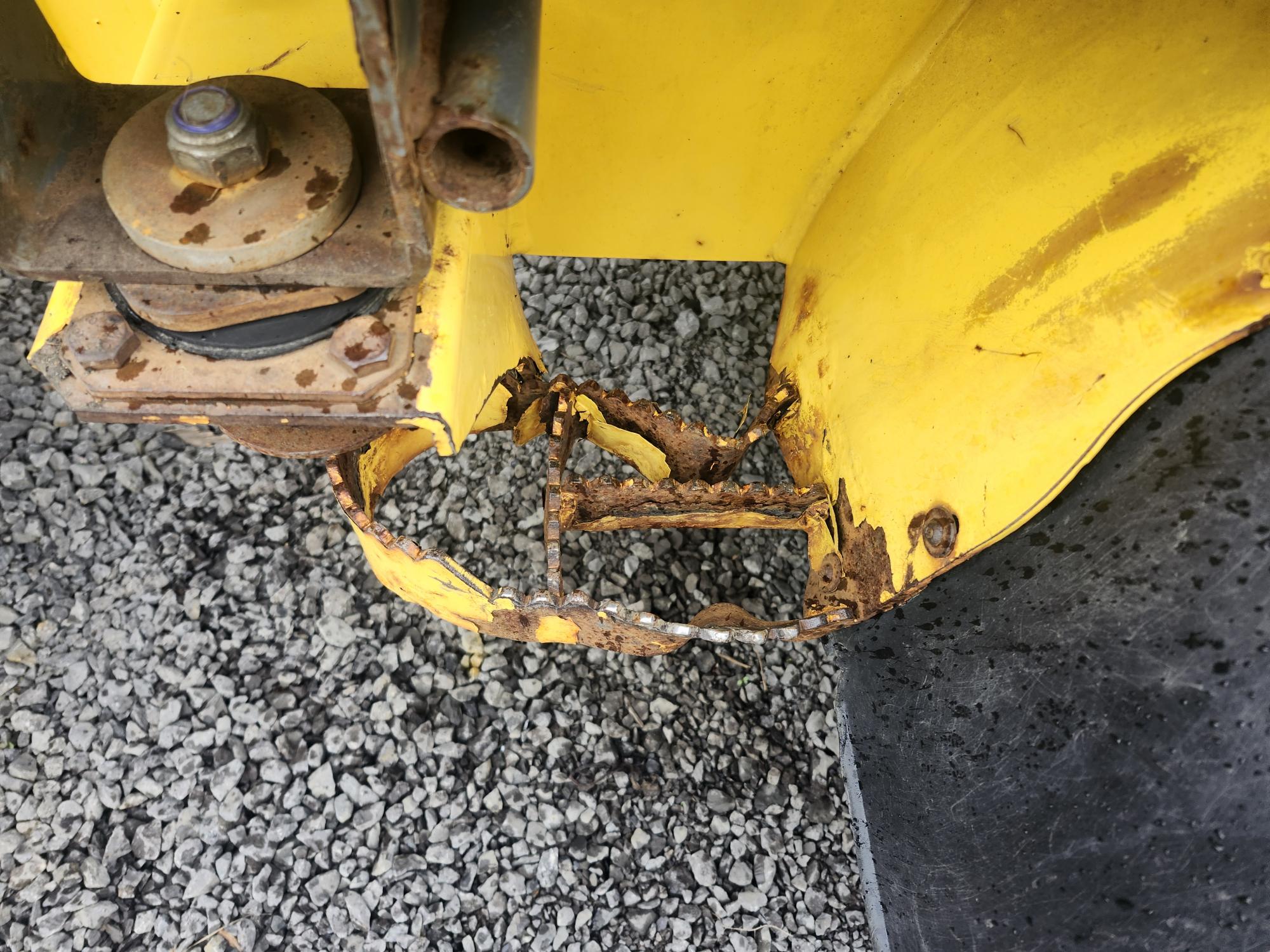 Gros plan sur un garde-boue en métal jaune d'un véhicule, présentant d'importants dommages dus à la rouille et à la corrosion. Le métal est irrégulier et partiellement manquant, avec du gravier visible sous la zone endommagée. Un pneu noir se trouve à côté du garde-boue.