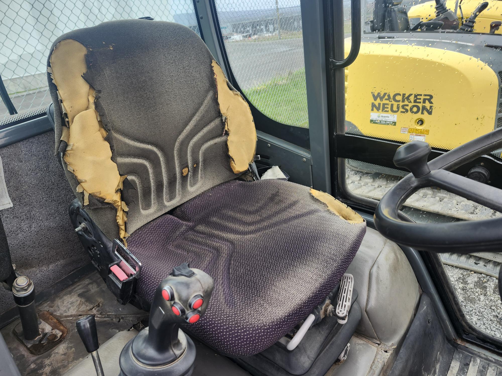 Un siège conducteur très usé dans un engin de chantier, dont les bords sont déchirés en mousse jaune. Un joystick et des commandes sont visibles, et une machine Wacker Neuson jaune est visible par la fenêtre.