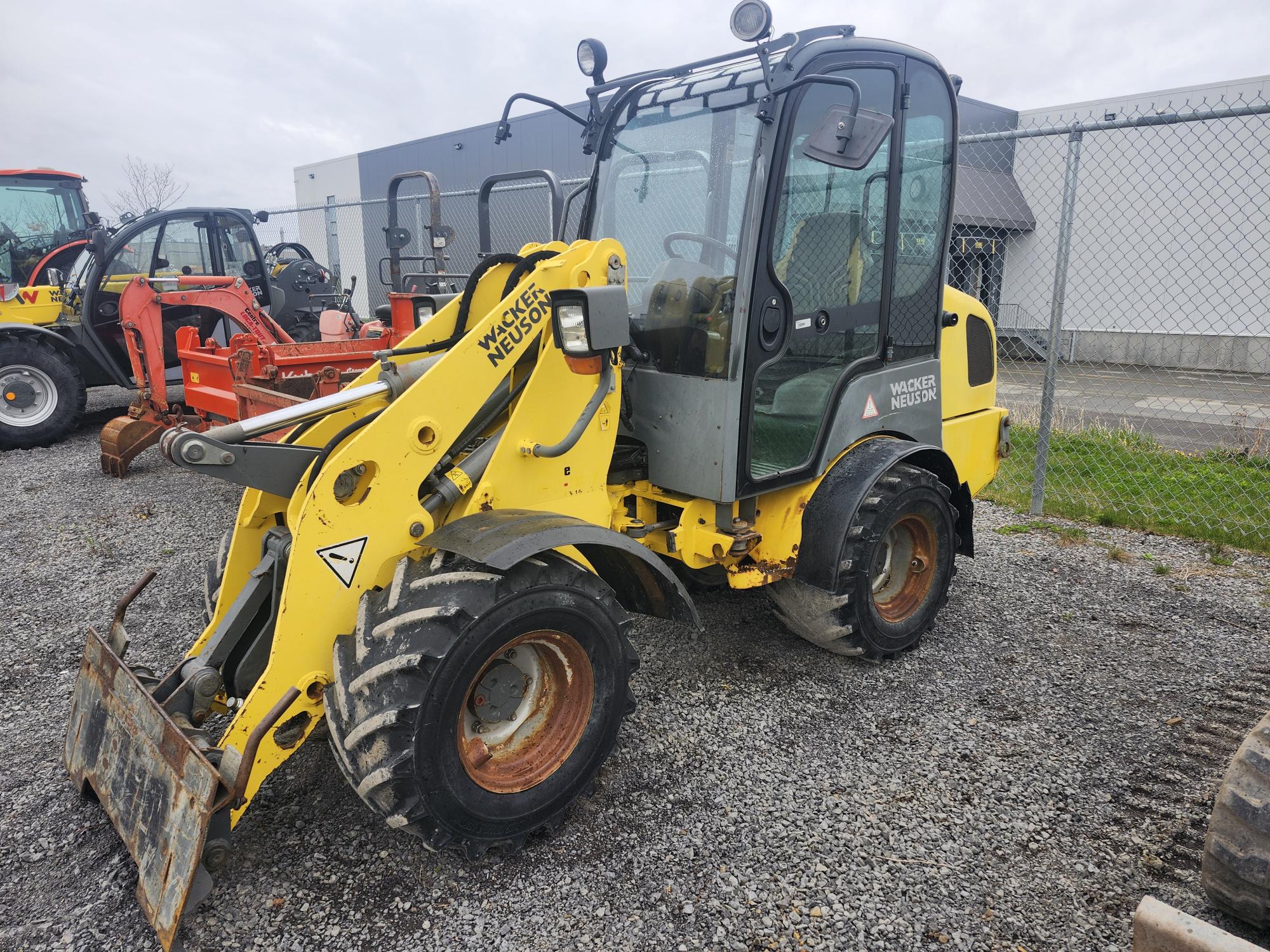 Une chargeuse sur pneus jaune Wacker Neuson est garée sur du gravier près d'une clôture grillagée, avec d'autres équipements de construction et un bâtiment gris en arrière-plan.