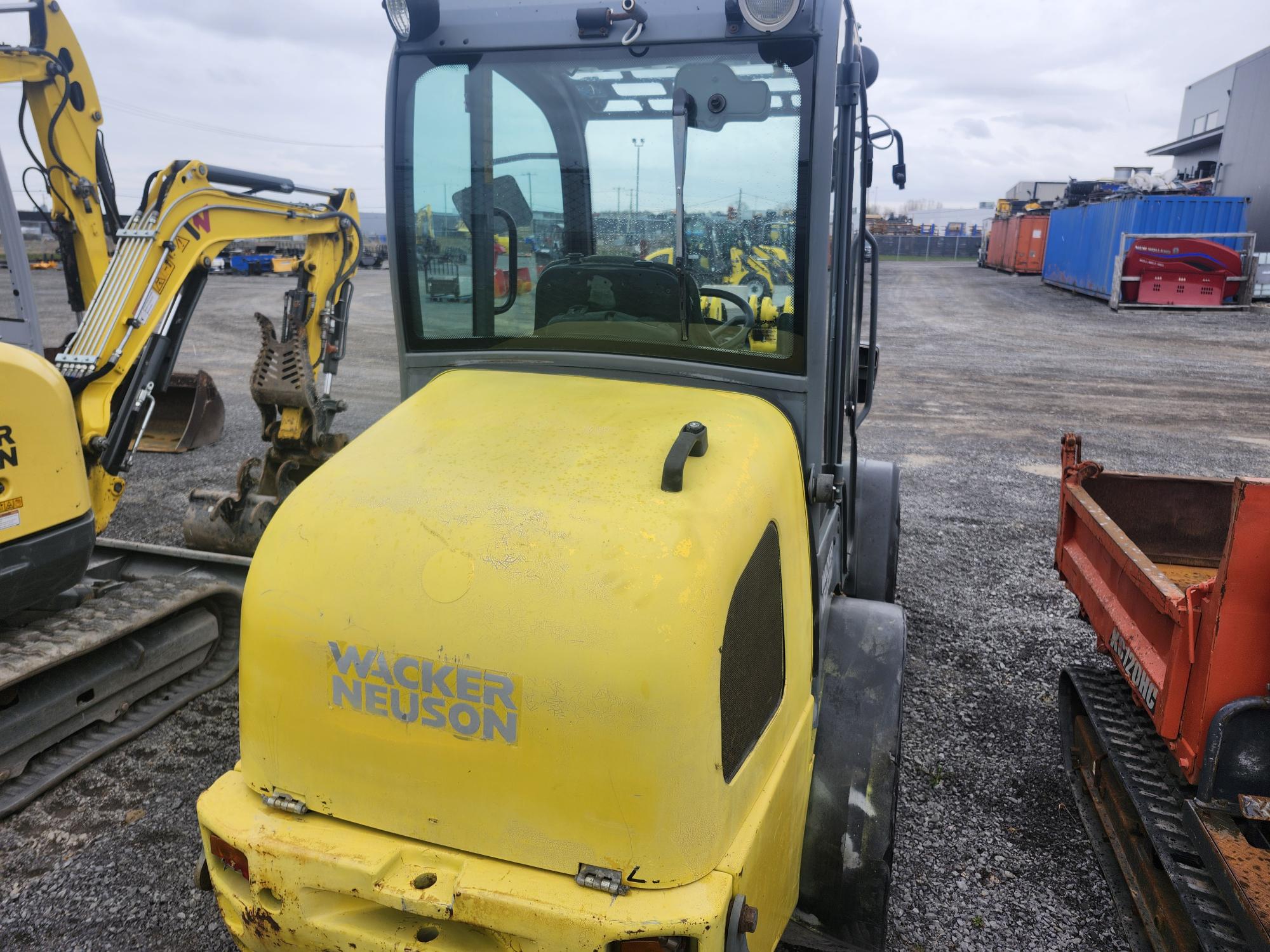 Un véhicule de construction jaune Wacker Neuson est garé sur du gravier à proximité d'autres machines, de conteneurs industriels et de bâtiments par une journée nuageuse.