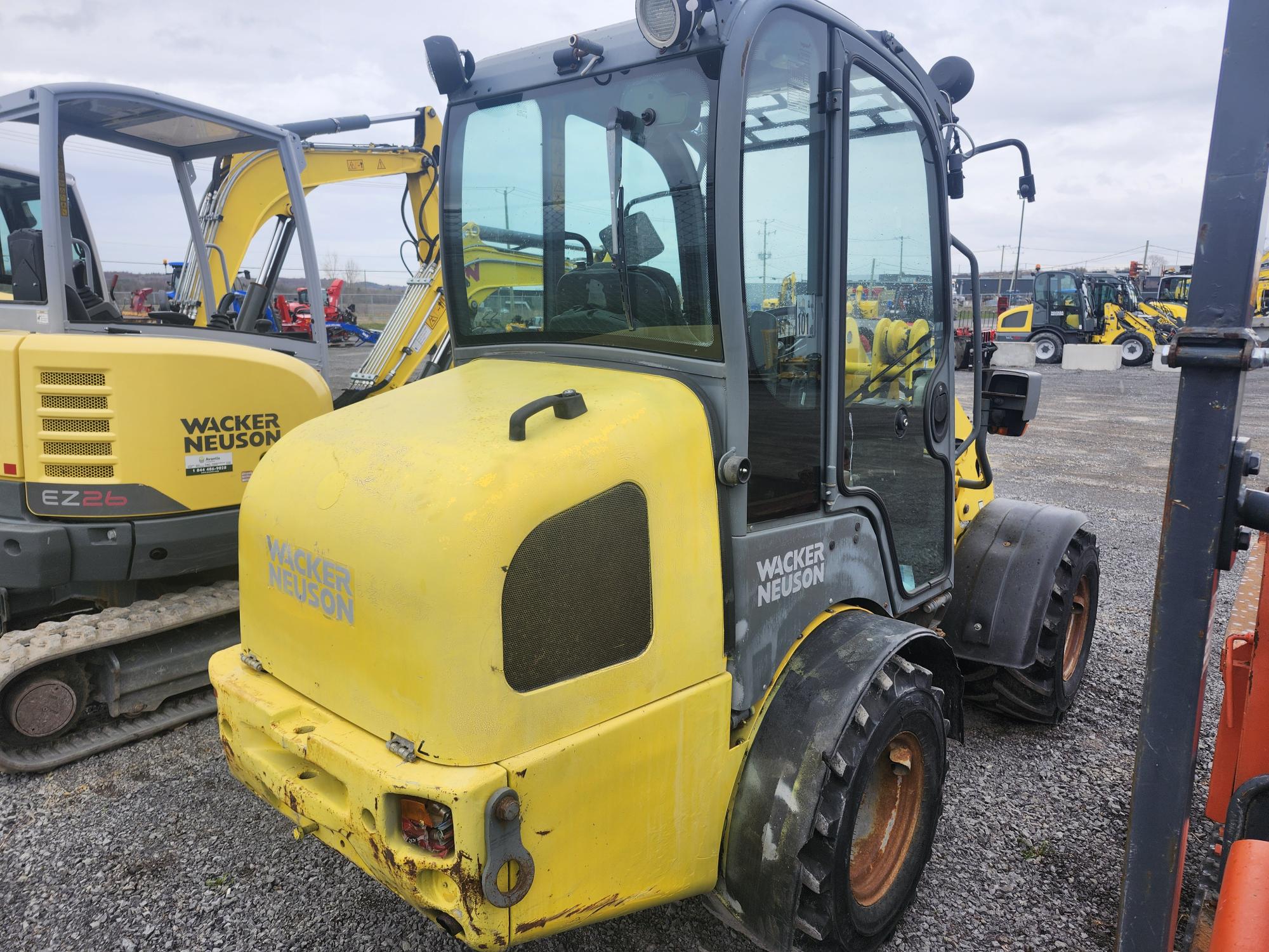 Un engin de chantier jaune Wacker Neuson est garé sur du gravier, à proximité d'autres engins. Il est équipé d'une cabine fermée, de gros pneus et est positionné face à la caméra.