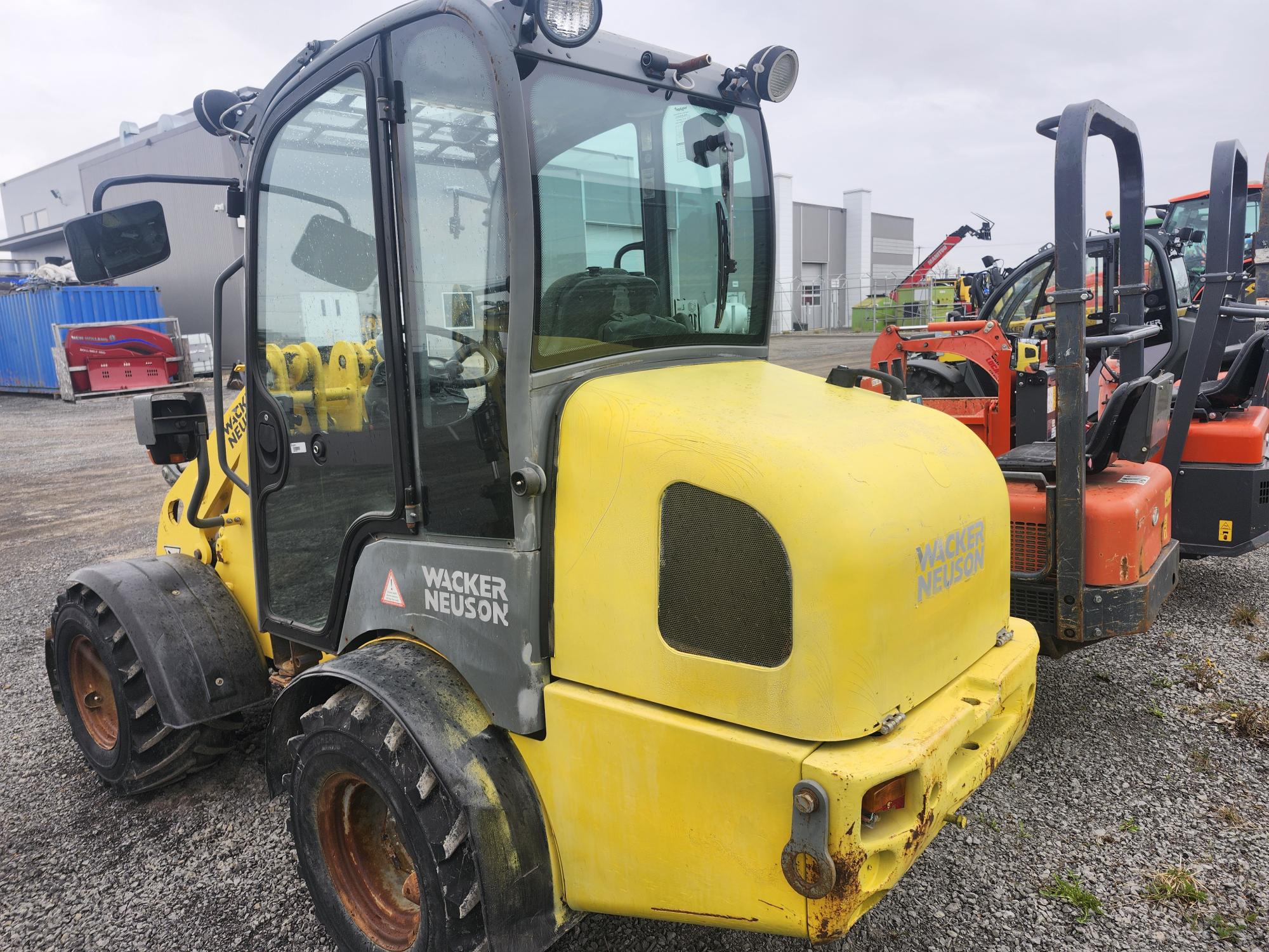 Un véhicule de construction jaune Wacker Neuson est garé sur du gravier à proximité d'autres machines orange, avec des bâtiments industriels visibles en arrière-plan sous un ciel nuageux.