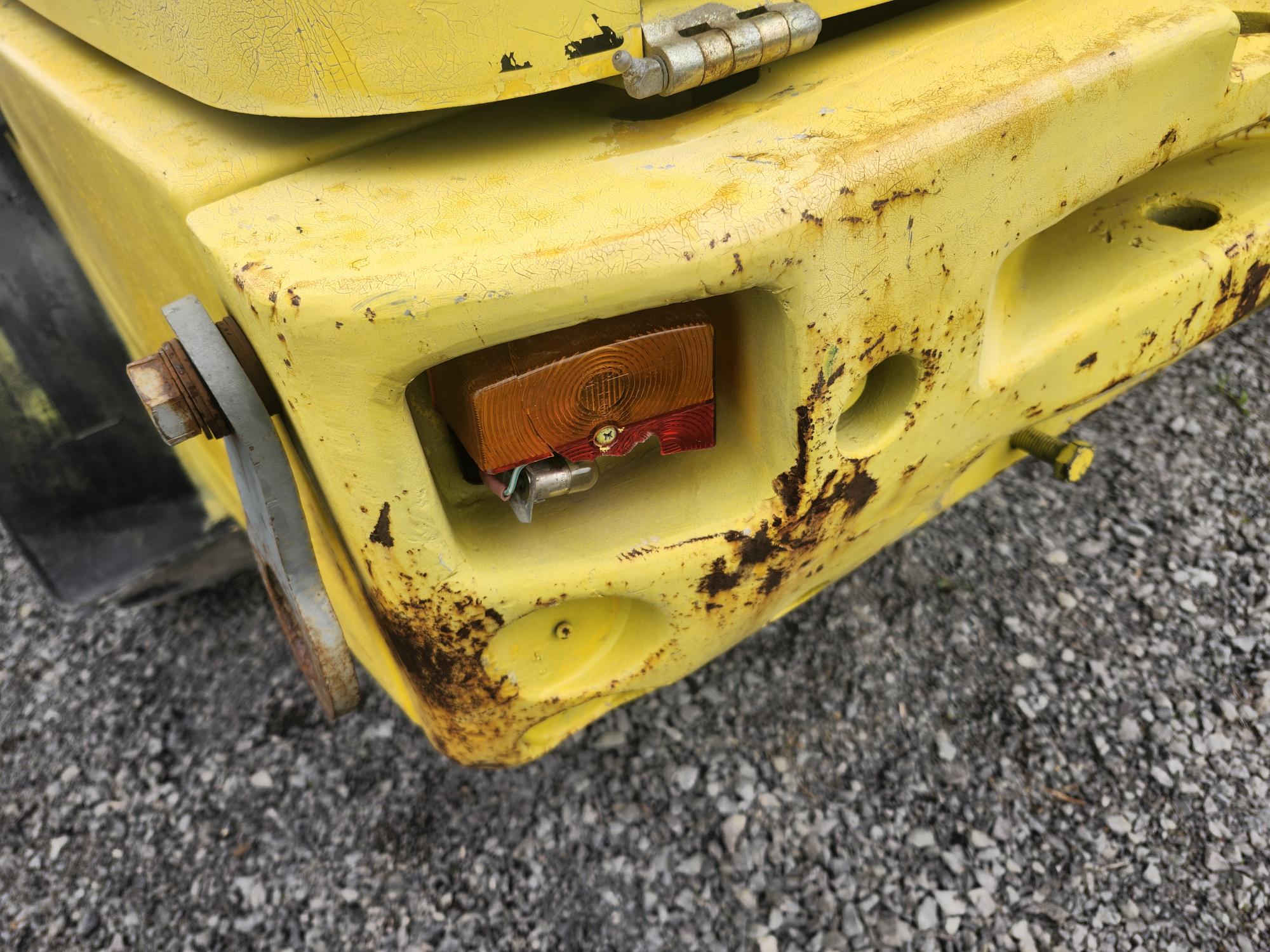 Gros plan d'une machine industrielle jaune patinée par le temps, avec des taches de rouille. Un feu arrière rouge et orange est visible, entouré de peinture écaillée, et la surface est rugueuse. La machine repose sur un lit de gravier gris.