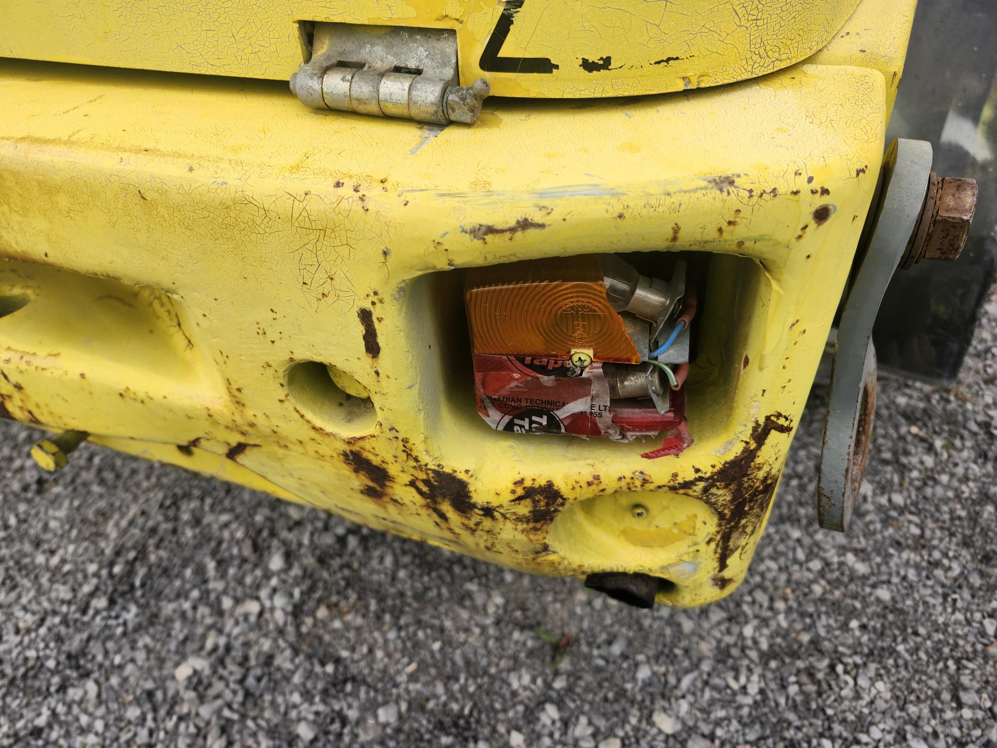 Gros plan sur un pare-chocs jaune usé, la peinture écaillée et rouillée ; un compartiment d'éclairage cassé contient des déchets, dont un emballage de snack froissé et de petits débris. Du gravier est visible au sol en contrebas.