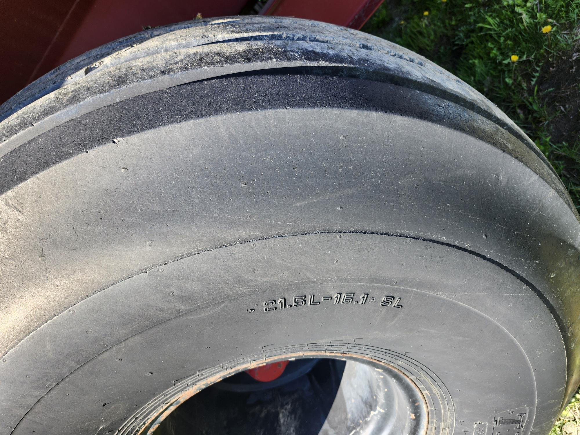 Gros plan d'un gros pneu usé à bande de roulement lisse, portant l'inscription « 21.5L-16.1 SL » sur le flanc. De l'herbe et une partie d'un véhicule rouge apparaissent en arrière-plan.