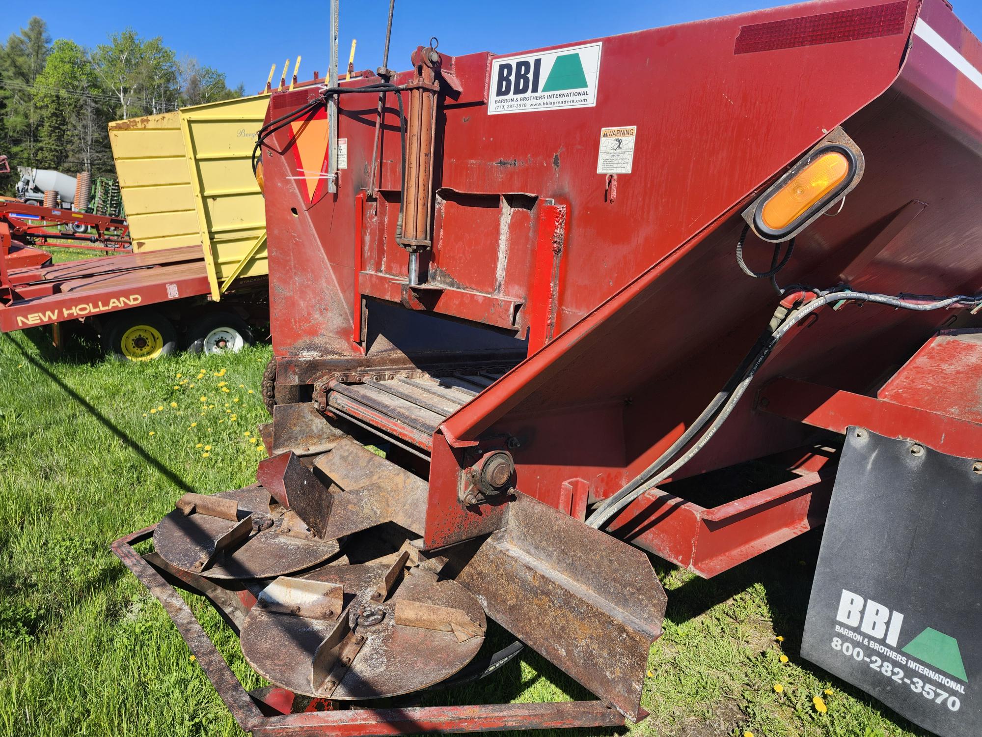 Gros plan d'une machine agricole rouge à lames métalliques rotatives, étiquetée « BBI ». Une autre machine New Holland jaune et rouge est visible à l'arrière-plan sur l'herbe verte sous un ciel bleu clair.