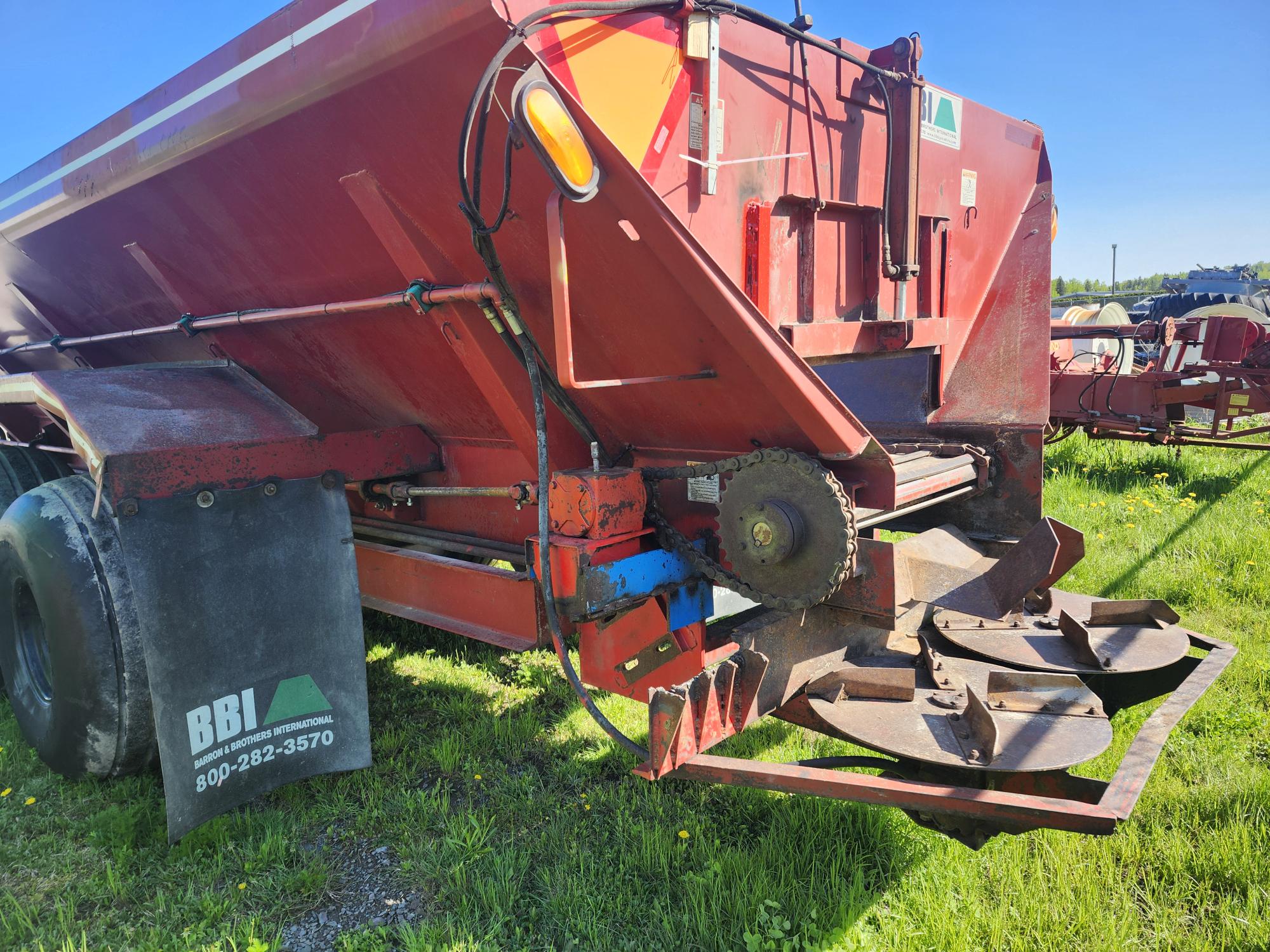 Remorque agricole rouge sur l'herbe verte, avec ses engrenages mécaniques, ses disques rotatifs et son garde-boue noir portant l'inscription « BBI » et un numéro de téléphone. Le ciel bleu ensoleillé laisse entrevoir d'autres équipements agricoles en arrière-plan.