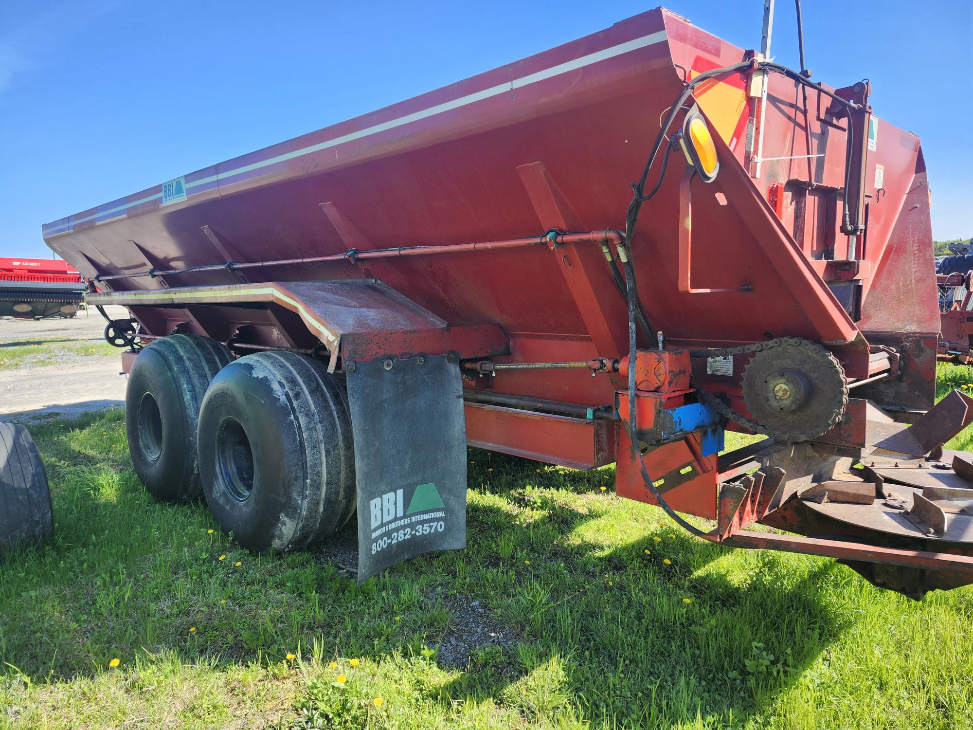 Une grande remorque agricole rouge à roues arrière jumelées est garée sur l'herbe. Elle est équipée de bavettes anti-boue étiquetées « BBI » et d'un mécanisme d'engrenage visible à l'avant. Le ciel bleu et d'autres équipements sont visibles en arrière-plan.