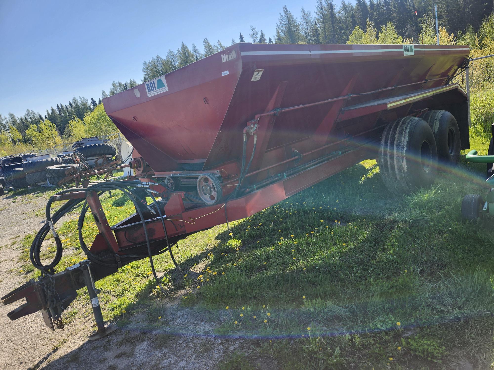 Une grande remorque agricole rouge à deux roues est garée sur l'herbe dans un espace extérieur ensoleillé, entourée d'arbres et de pièces de machines. Un léger halo arc-en-ciel est visible sur l'image.