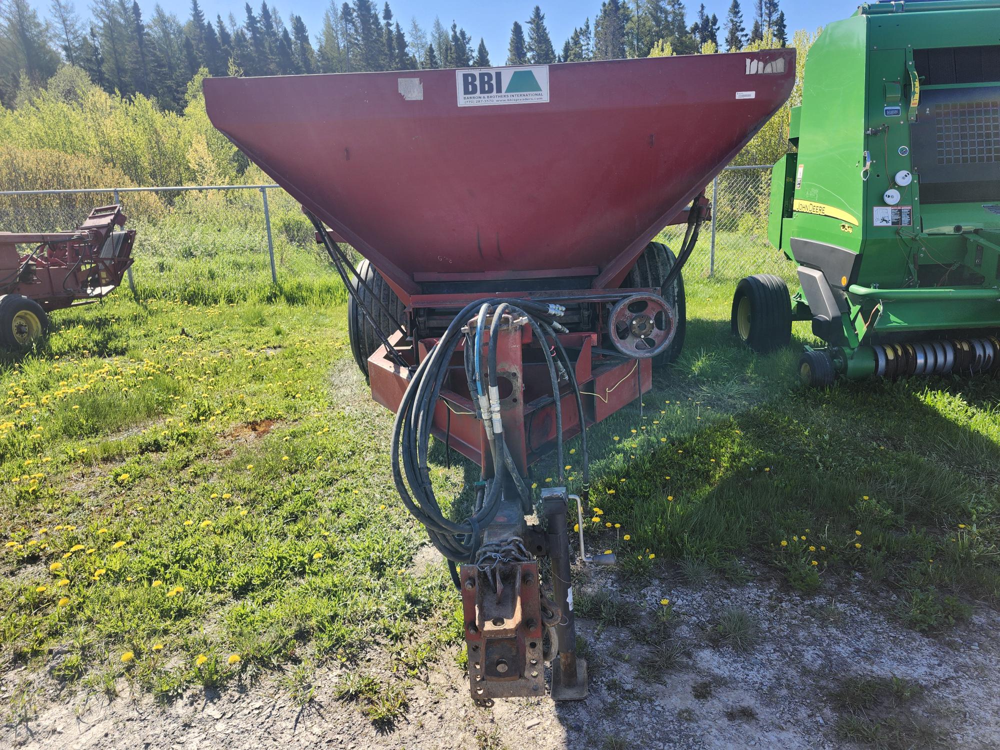 Un épandeur agricole BBI rouge est posé sur l'herbe, avec des flexibles hydrauliques et des pièces métalliques visibles à l'avant. Une machine agricole verte est partiellement visible à droite, avec des arbres et une clôture en fil de fer en arrière-plan.