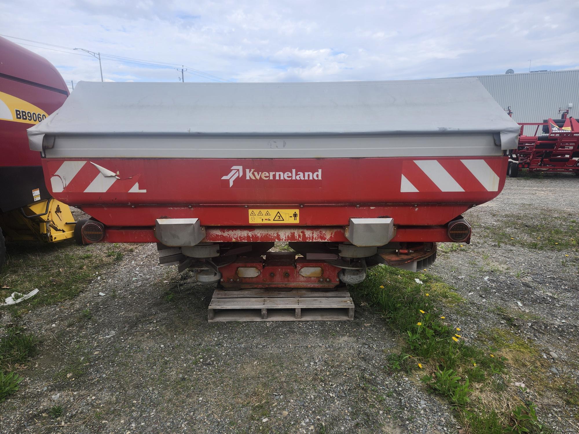 Une machine agricole Kverneland rouge avec un toit gris et des étiquettes d'avertissement est garée à l'extérieur sur du gravier, à proximité d'autres équipements agricoles.