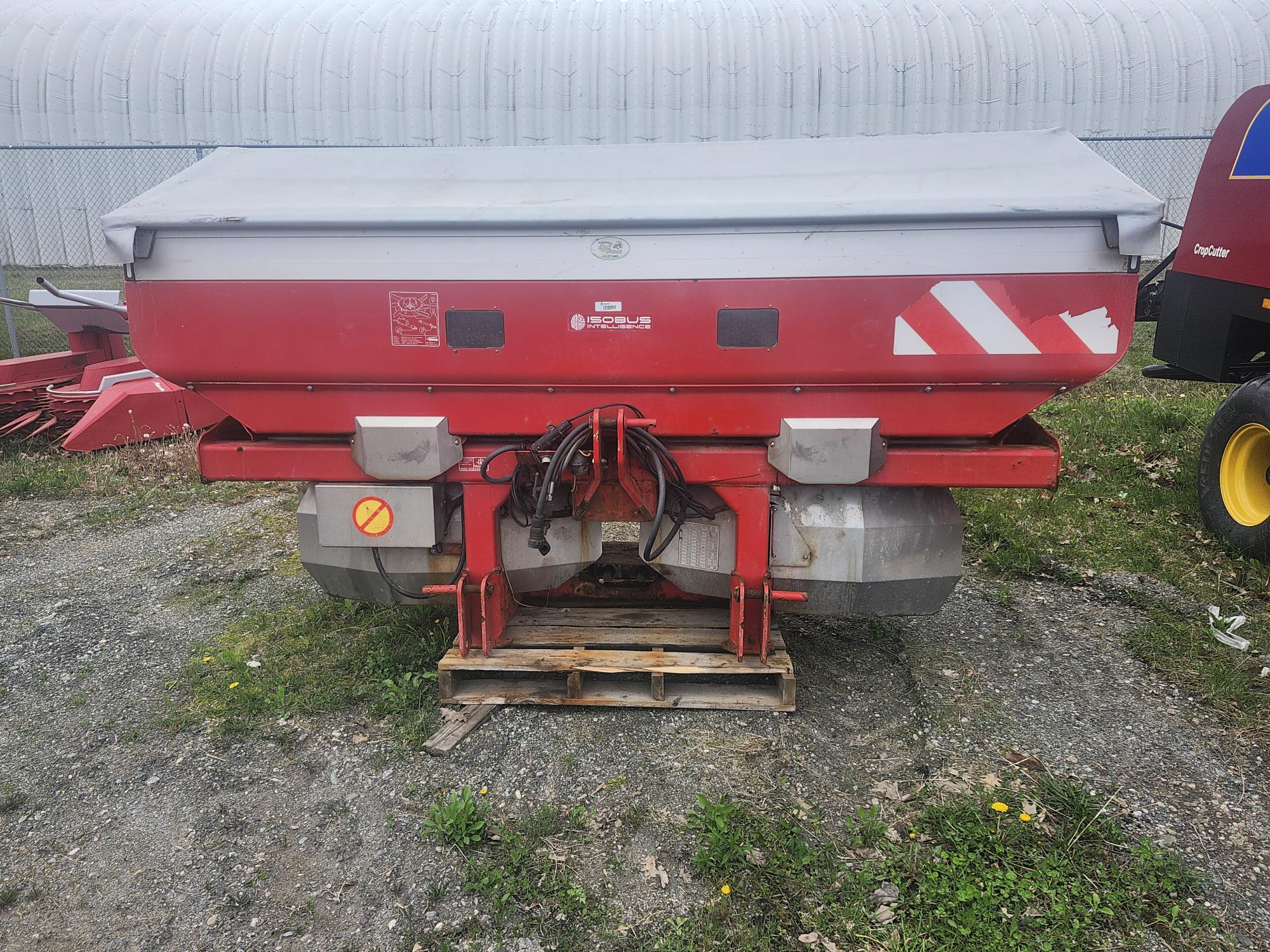 Une grande machine agricole rouge, probablement un épandeur d'engrais, est posée sur une palette en bois à l'extérieur. L'épandeur est équipé d'un capot gris et de divers tuyaux et accessoires, avec un bâtiment métallique visible en arrière-plan.