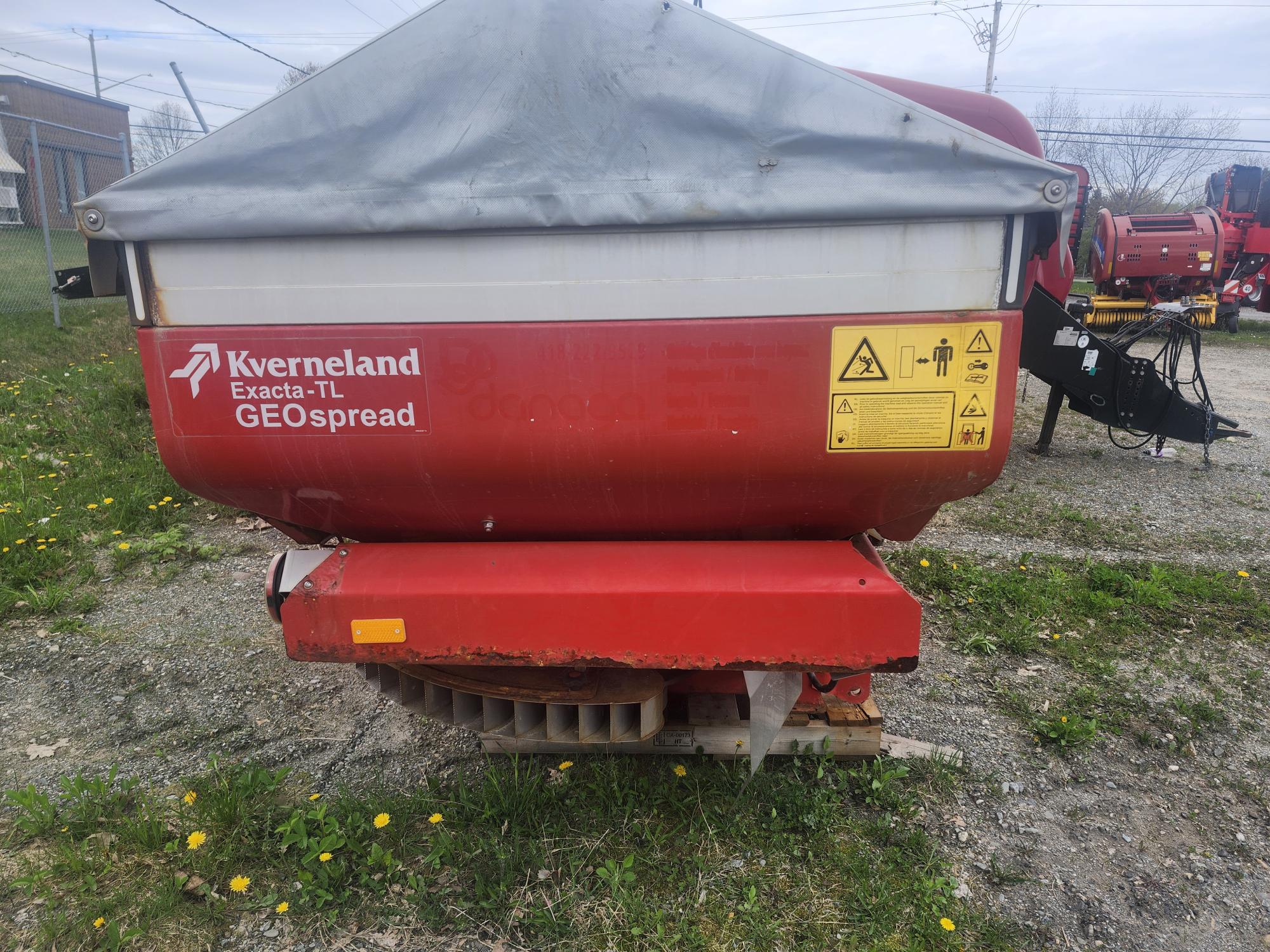 Une machine agricole Kverneland Exacta-TL GEOspread rouge repose sur du gravier et de l'herbe, avec des étiquettes d'avertissement et une bâche grise. Des pissenlits jaunes et d'autres machines sont visibles en arrière-plan.