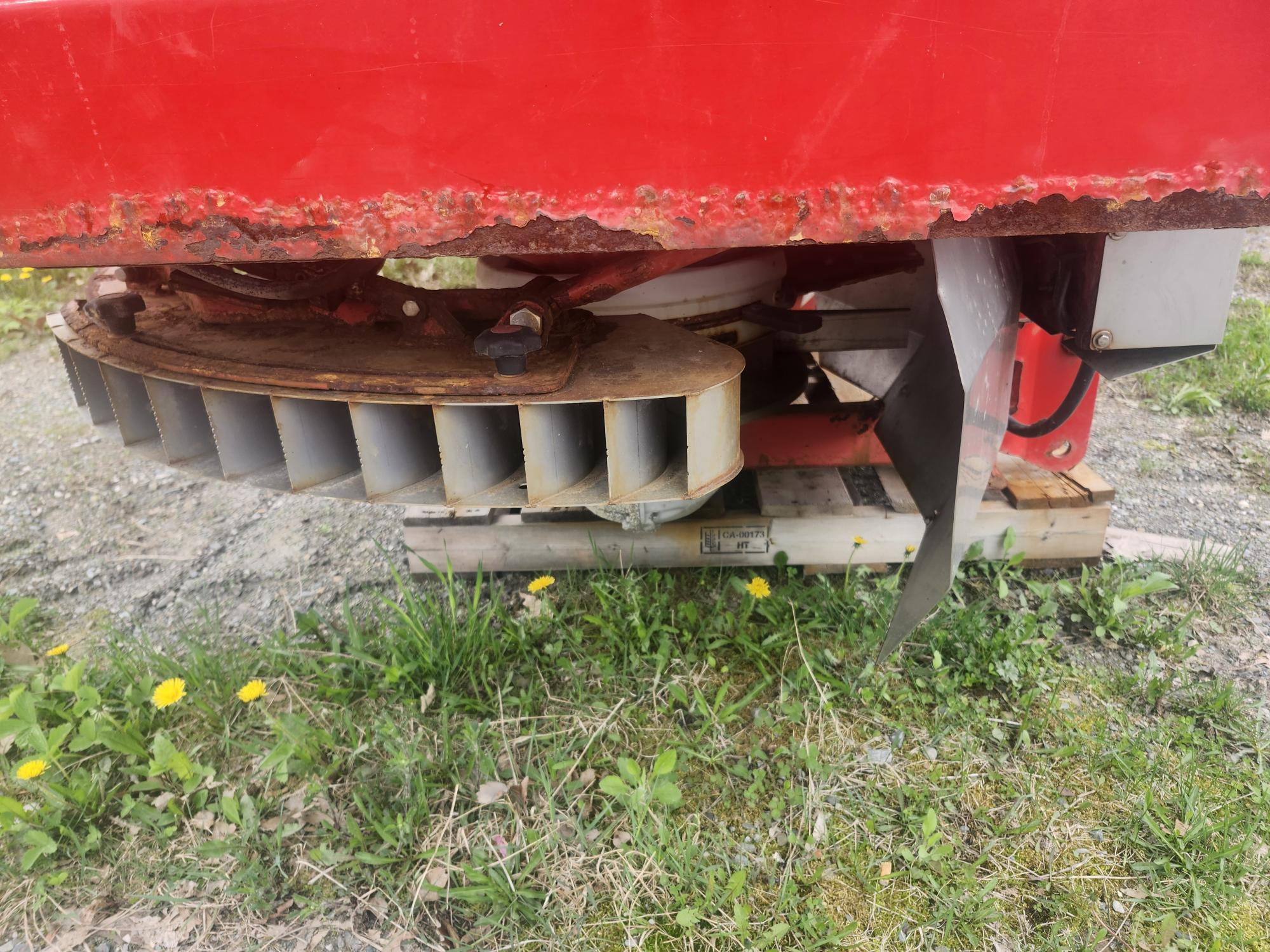 Gros plan du dessous d'une machine en métal rouge avec de la rouille visible, montrant une roue métallique segmentée et une fixation de lame, posée sur une palette en bois à l'extérieur sur de l'herbe et du gravier avec des pissenlits jaunes.