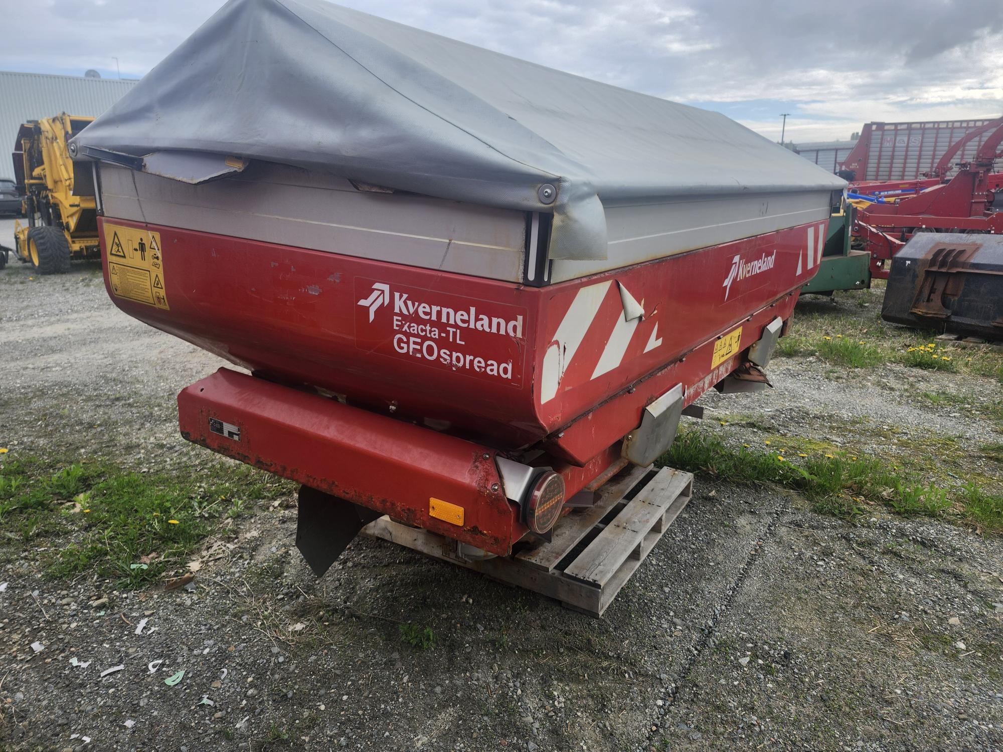 Une machine agricole Kverneland Exacta-TL GEOspread rouge avec une couverture grise est placée sur des palettes en bois à l'extérieur, entourée de gravier et d'autres équipements agricoles.