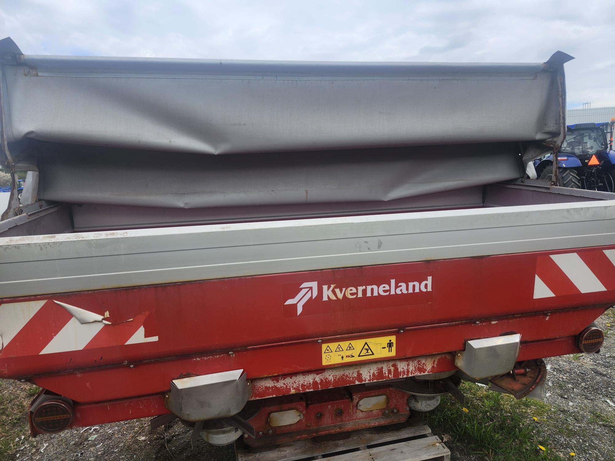 Une machine agricole Kverneland rouge et argent, avec toit couvert, posée sur un sol irrégulier. Des étiquettes d'avertissement et des bandes réfléchissantes sont visibles sur son panneau avant.