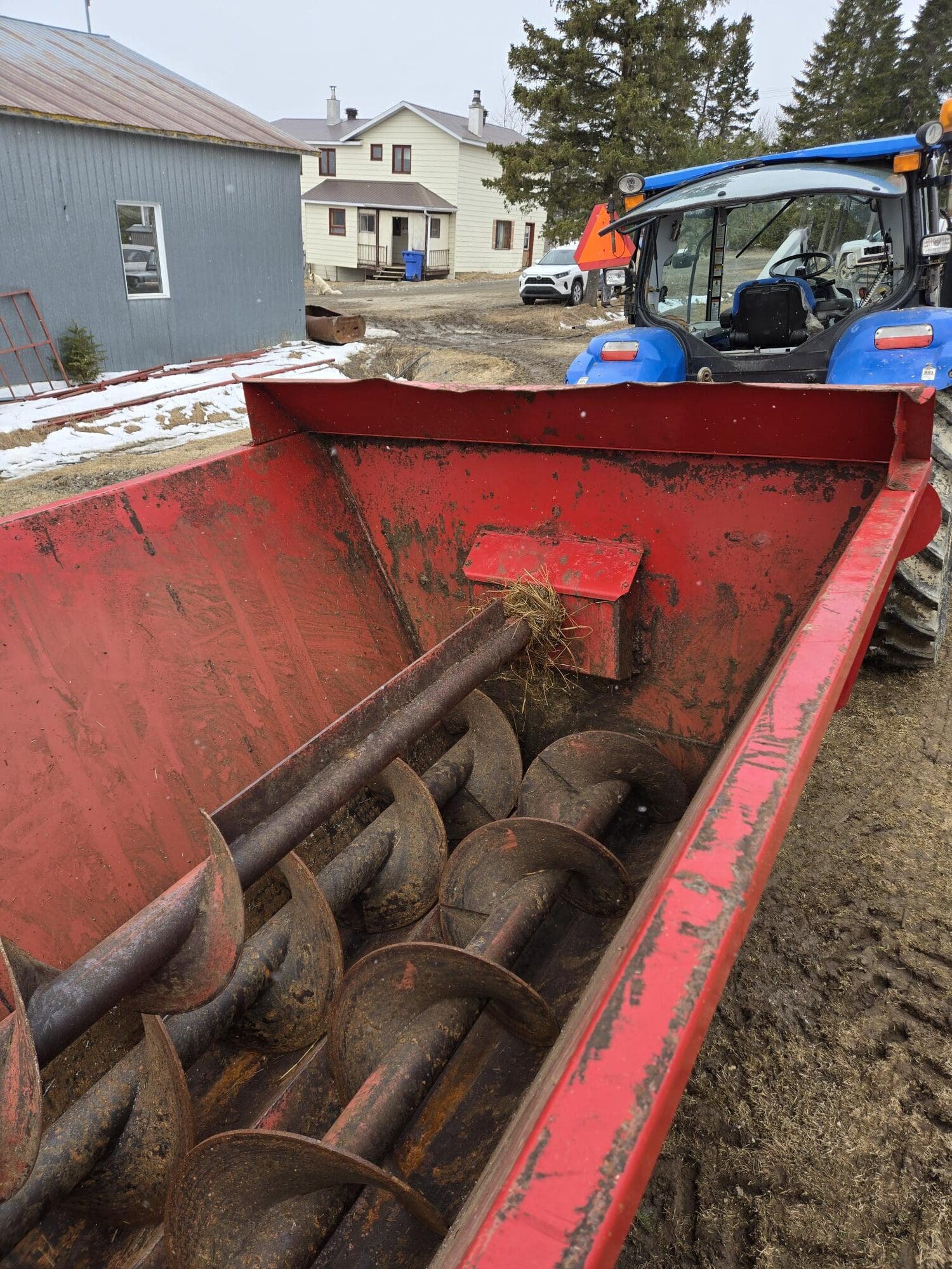 Vue de derrière un mélangeur d'aliments rouge avec des vis sans fin, attaché à un tracteur bleu garé dans une cour de ferme boueuse ; des bâtiments et des arbres sont en arrière-plan, avec quelques plaques de neige visibles.