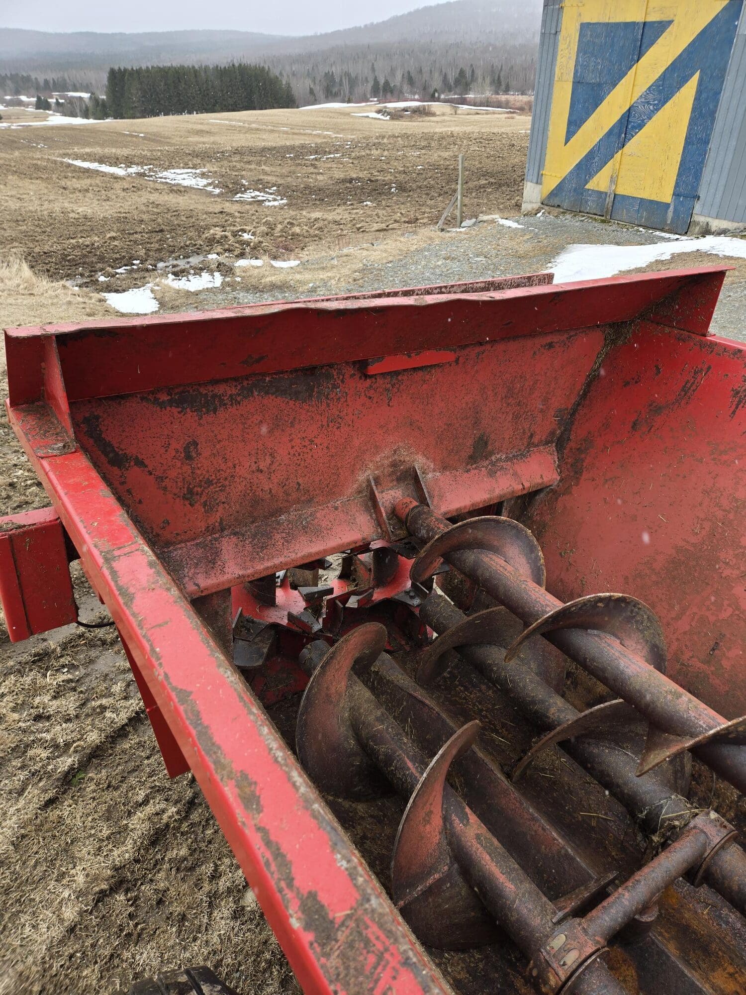 Gros plan d'une machine agricole rouge équipée de grandes vis sans fin, posée à l'extérieur sur un champ boueux parsemé de plaques de neige, avec un bâtiment bleu et jaune en arrière-plan. Des collines boisées sont visibles au loin.