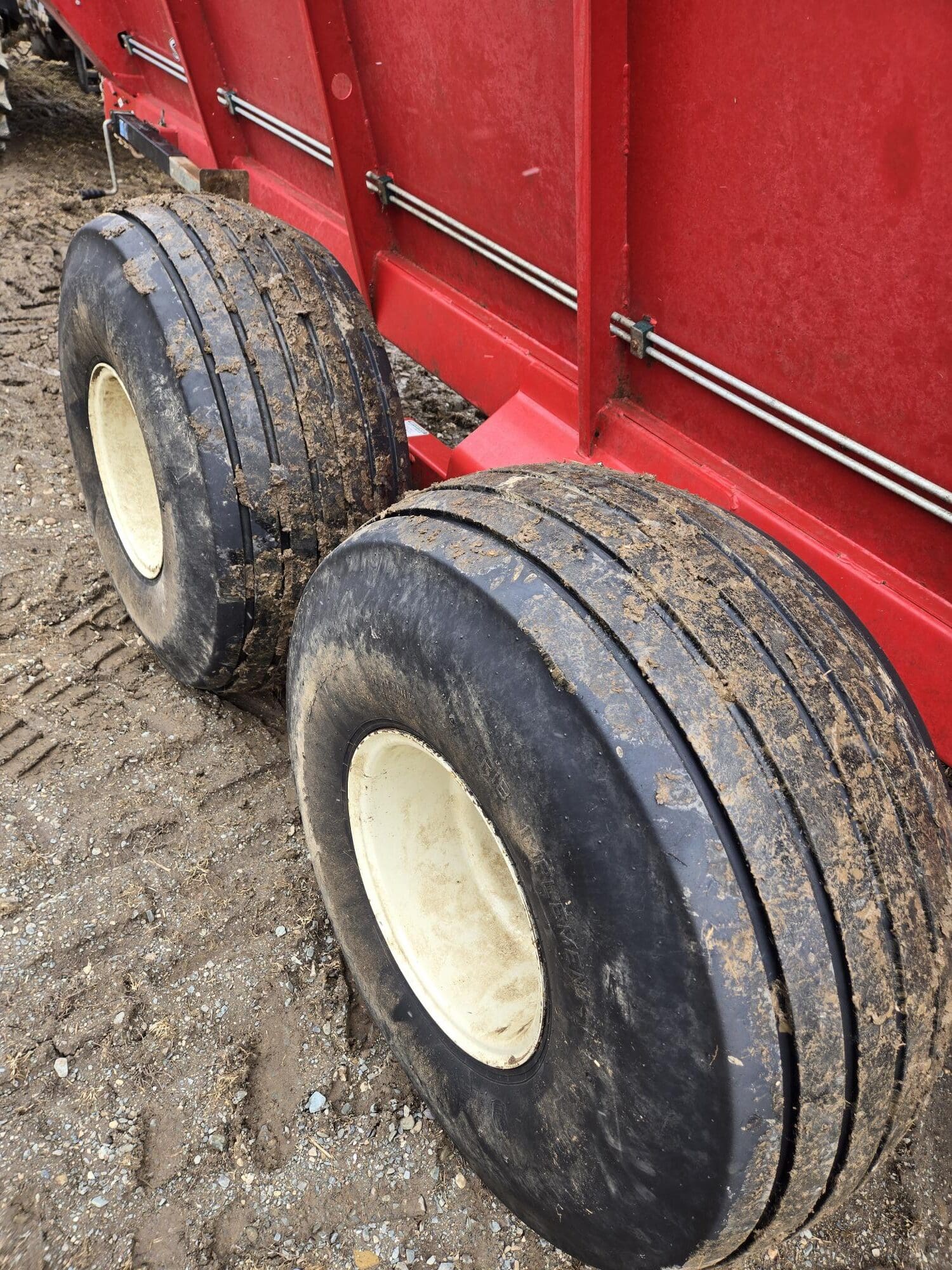 Gros plan de deux gros pneus boueux avec des jantes blanches attachés à une remorque ou une machine agricole rouge, garée sur une surface de terre et de gravier.