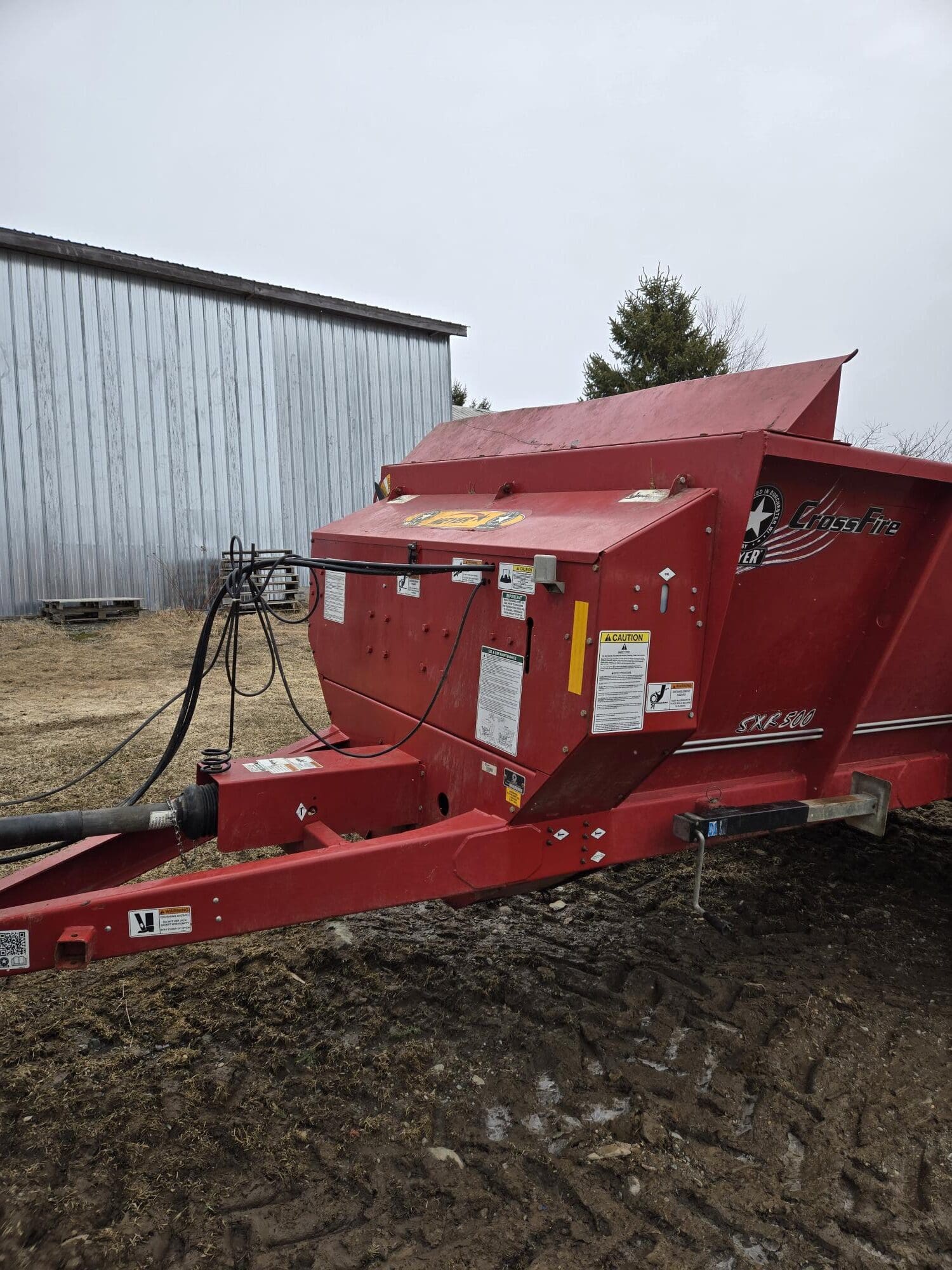 Une grande machine agricole rouge, probablement un mélangeur ou un broyeur d'aliments pour animaux, repose sur un sol boueux à côté d'une grange aux parois métalliques. Diverses étiquettes d'avertissement et câbles sont visibles sur la machine. Des arbres apparaissent en arrière-plan.