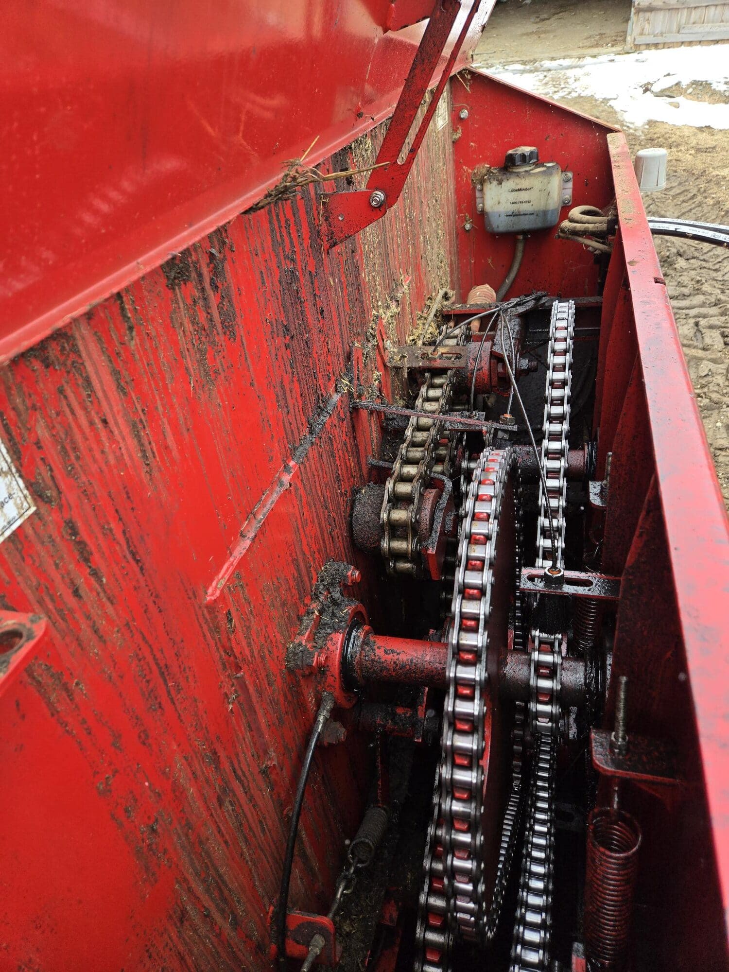 Gros plan de l'intérieur d'une machine agricole rouge montrant plusieurs engrenages et chaînes, avec de la terre et de la paille autour des mécanismes. Un récipient contenant du liquide est fixé en haut à droite.