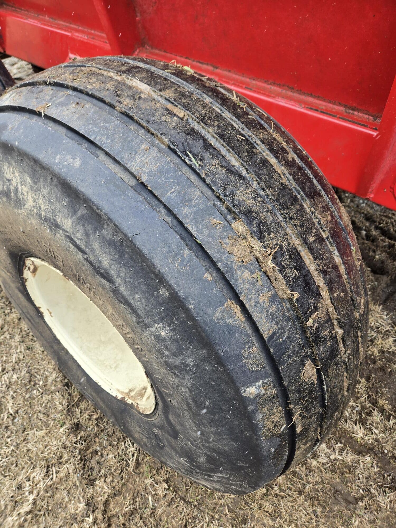 Gros plan d'un gros pneu en caoutchouc noir avec trois rainures de bande de roulement, recouvert de terre et de brins d'herbe, attaché à une machine ou un équipement rouge sur une surface herbeuse sèche.