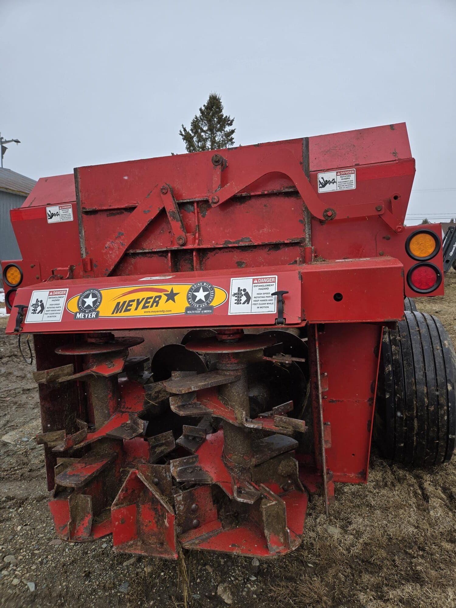 Une machine agricole rouge Meyer est posée sur la terre, affichant de grandes lames rotatives et plusieurs étiquettes d'avertissement à l'arrière, avec une paire de grands pneus noirs à droite et un arbre en arrière-plan.
