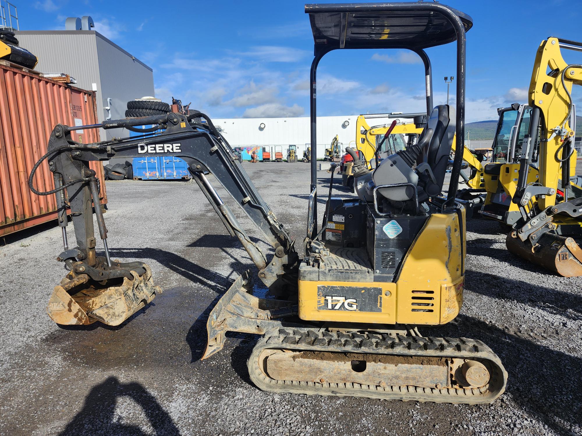 Une mini-pelle John Deere 17G jaune et noire avec chenilles en caoutchouc est garée sur du gravier à proximité d'autres équipements de construction et de conteneurs de fret sous un ciel bleu.