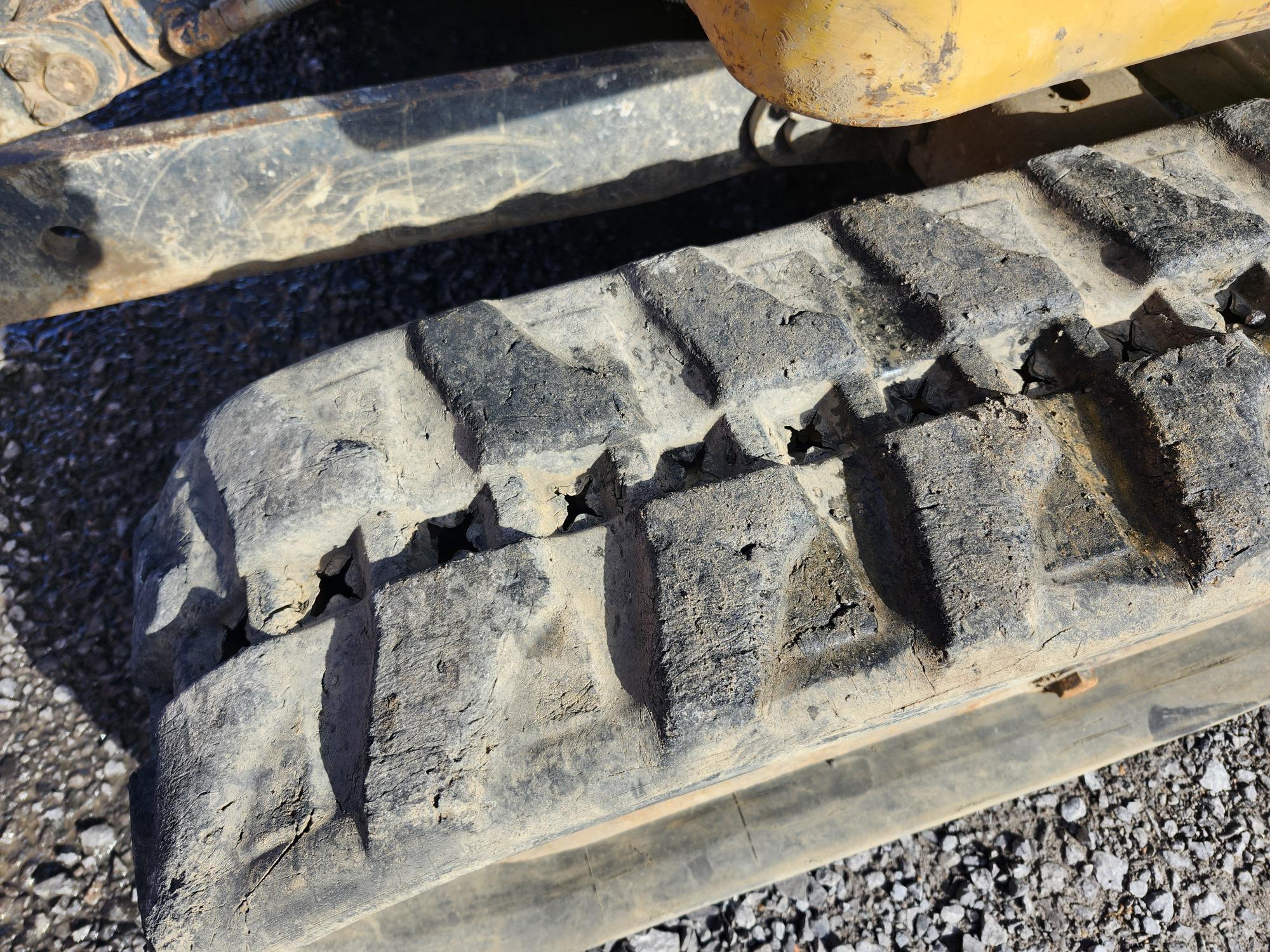 Gros plan d'une chenille en caoutchouc sale et usée sur un engin de chantier, avec du gravier et une partie de la machine jaune visibles en arrière-plan.
