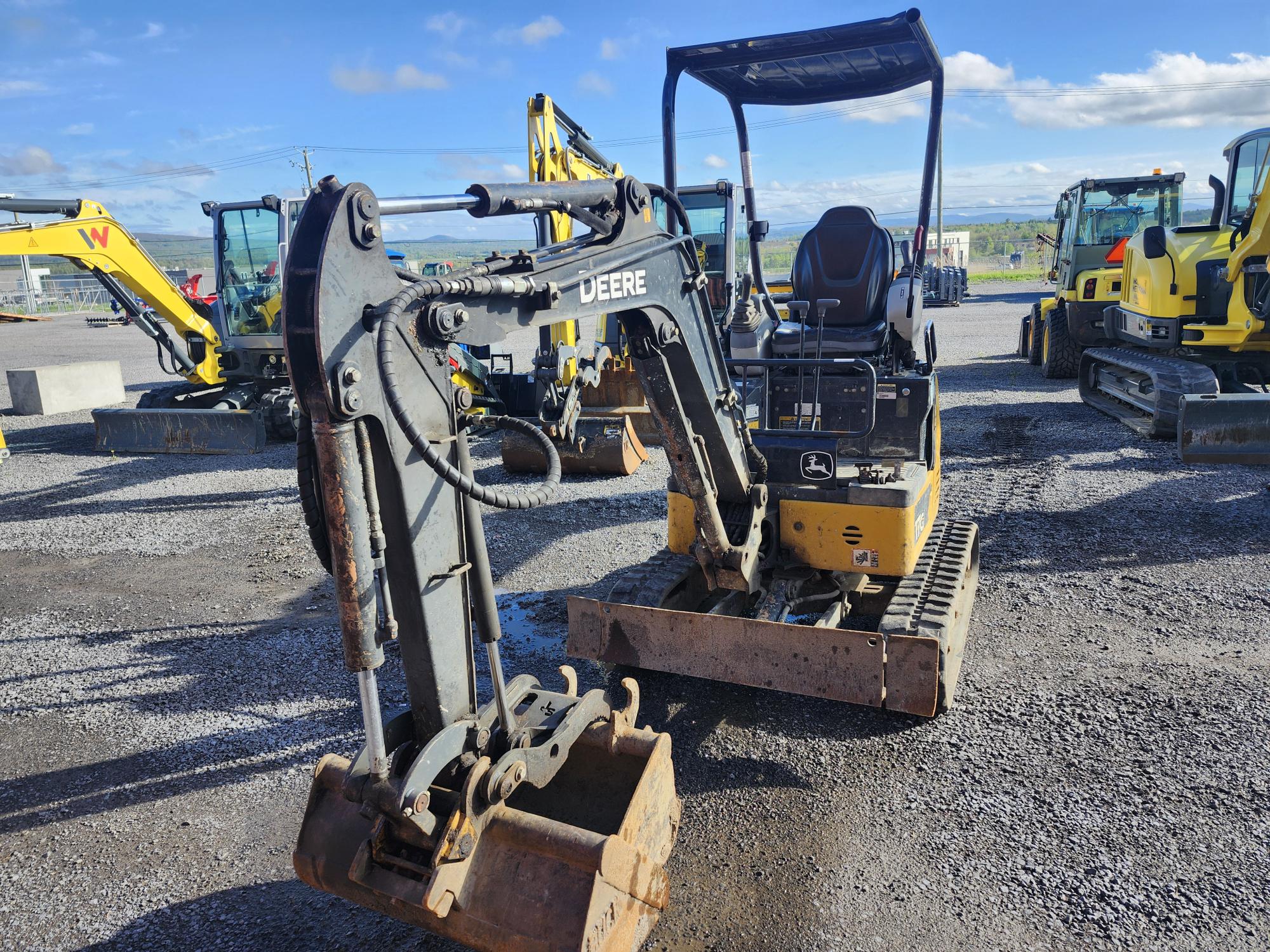 Une mini-pelle compacte jaune et noire John Deere est garée sur du gravier, avec d'autres véhicules de construction en arrière-plan sous un ciel bleu vif avec des nuages épars.
