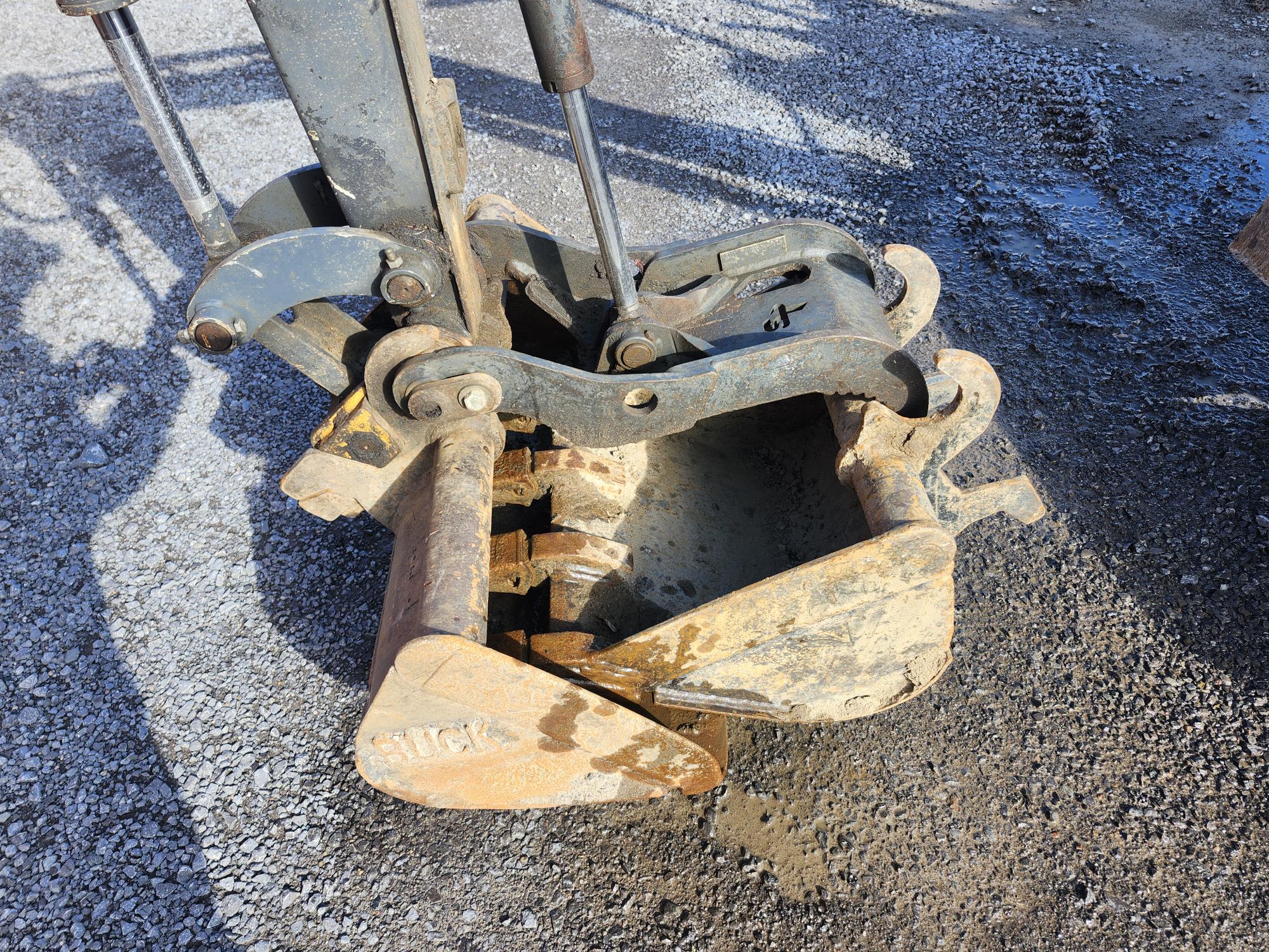 Gros plan d'un godet d'excavatrice en métal reposant sur du gravier, montrant des composants hydrauliques et des joints avec de la saleté et de la rouille visibles sur sa surface sous la lumière du soleil.