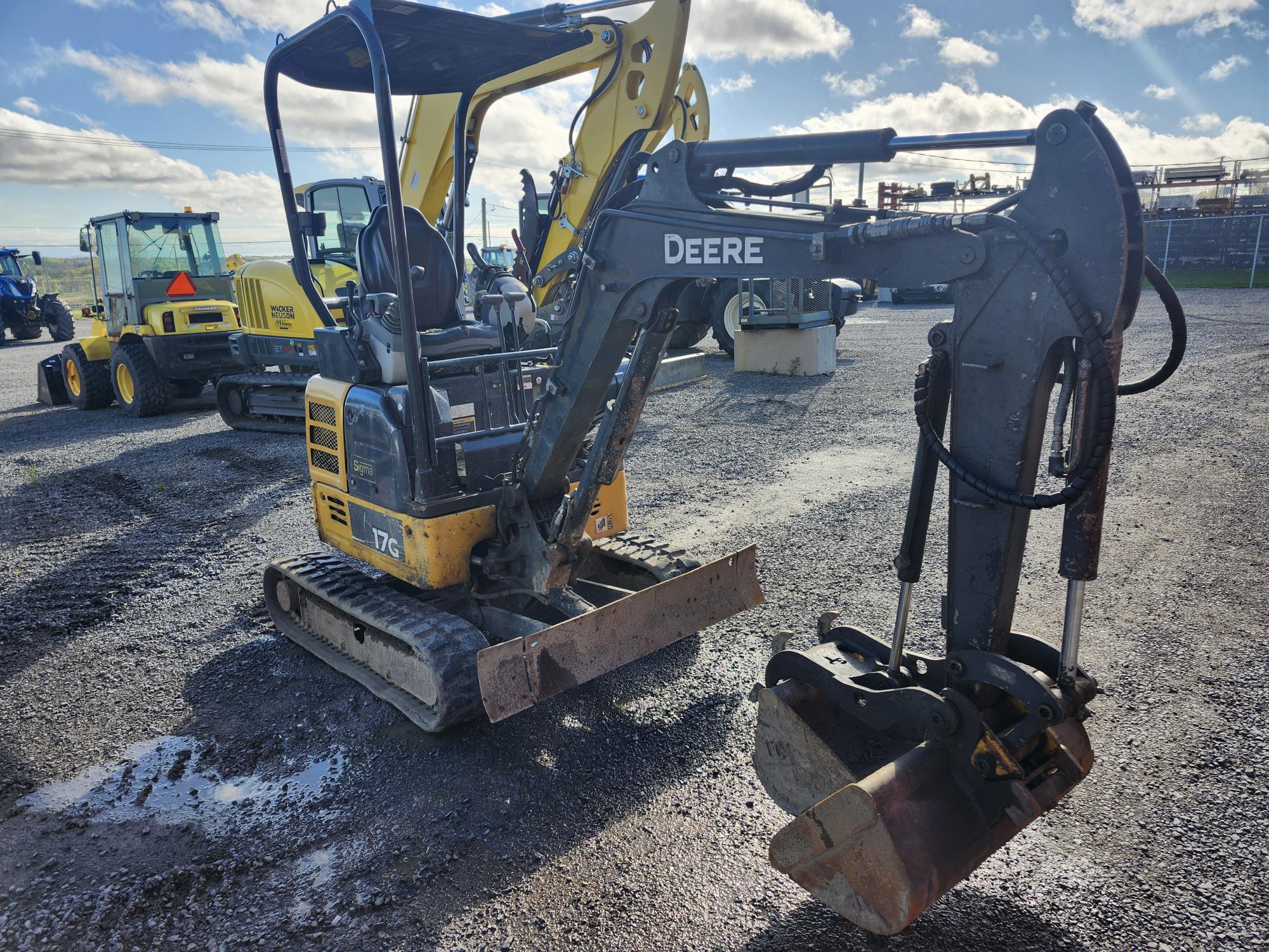 Une mini-pelle compacte John Deere avec un godet de creusement est garée sur un terrain en gravier sous un ciel partiellement nuageux, avec d'autres équipements de construction visibles en arrière-plan.