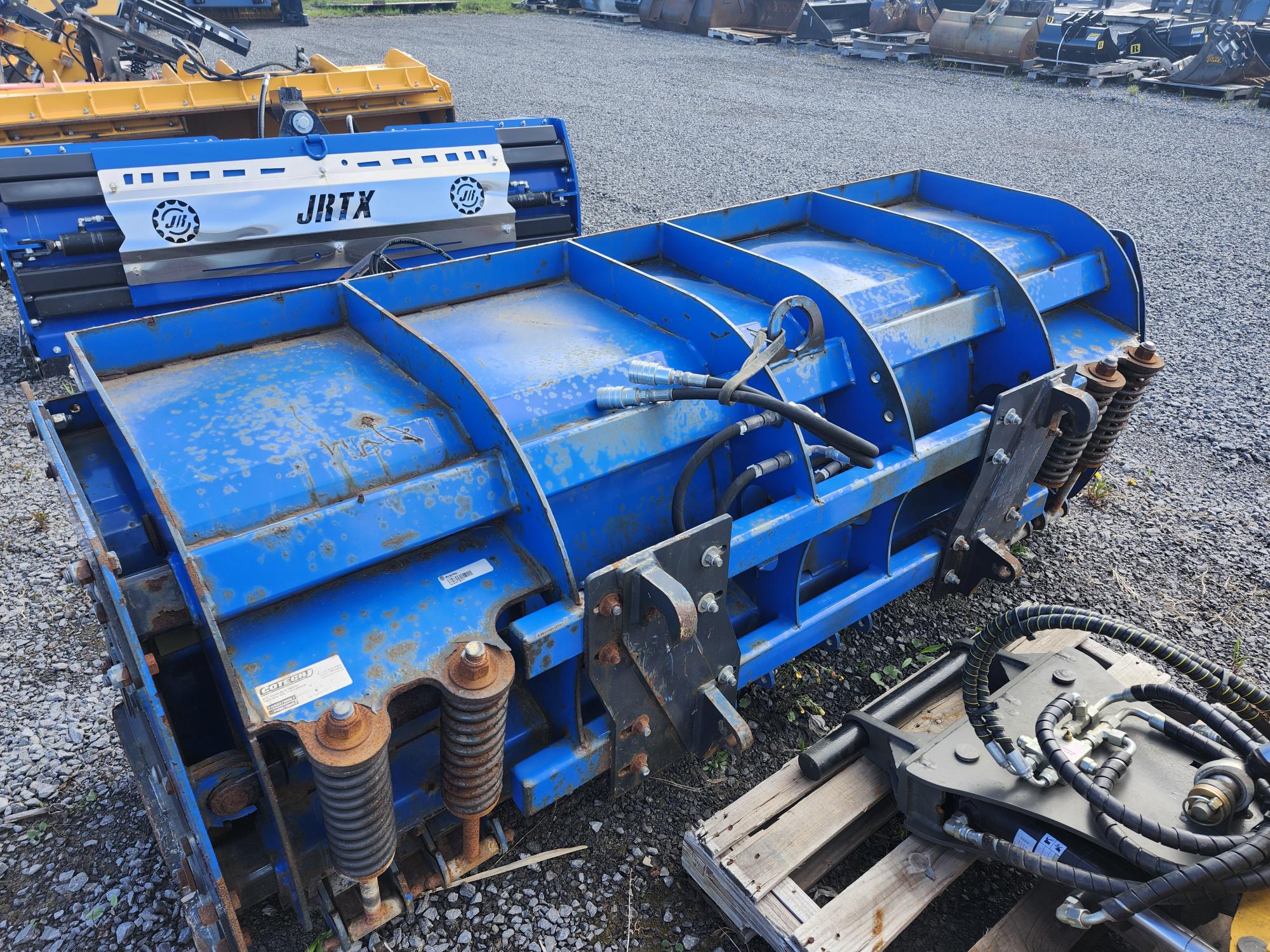 Un grand accessoire industriel bleu avec des taches de rouille, des tuyaux hydrauliques et des ressorts métalliques repose sur du gravier à côté d'autres pièces de machines et de palettes en bois.
