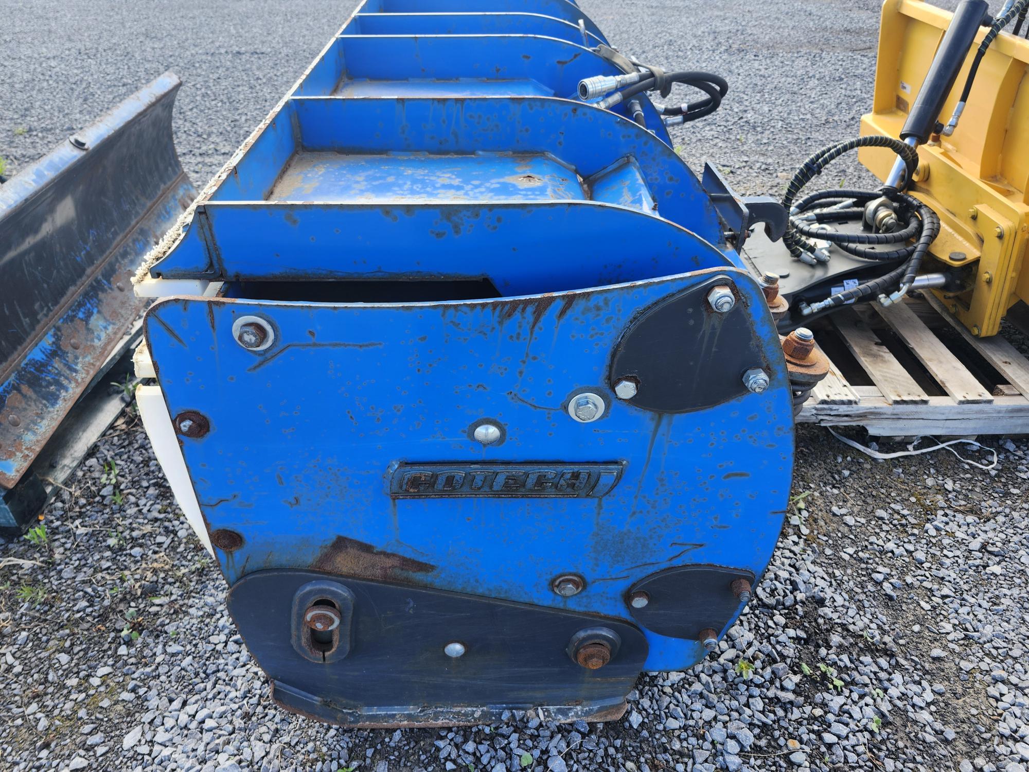 Gros plan d'un accessoire industriel en métal bleu, étiqueté « CMTBKI », posé sur du gravier, à proximité d'autres pièces et équipements. La surface présente des signes d'usure et de rouille.