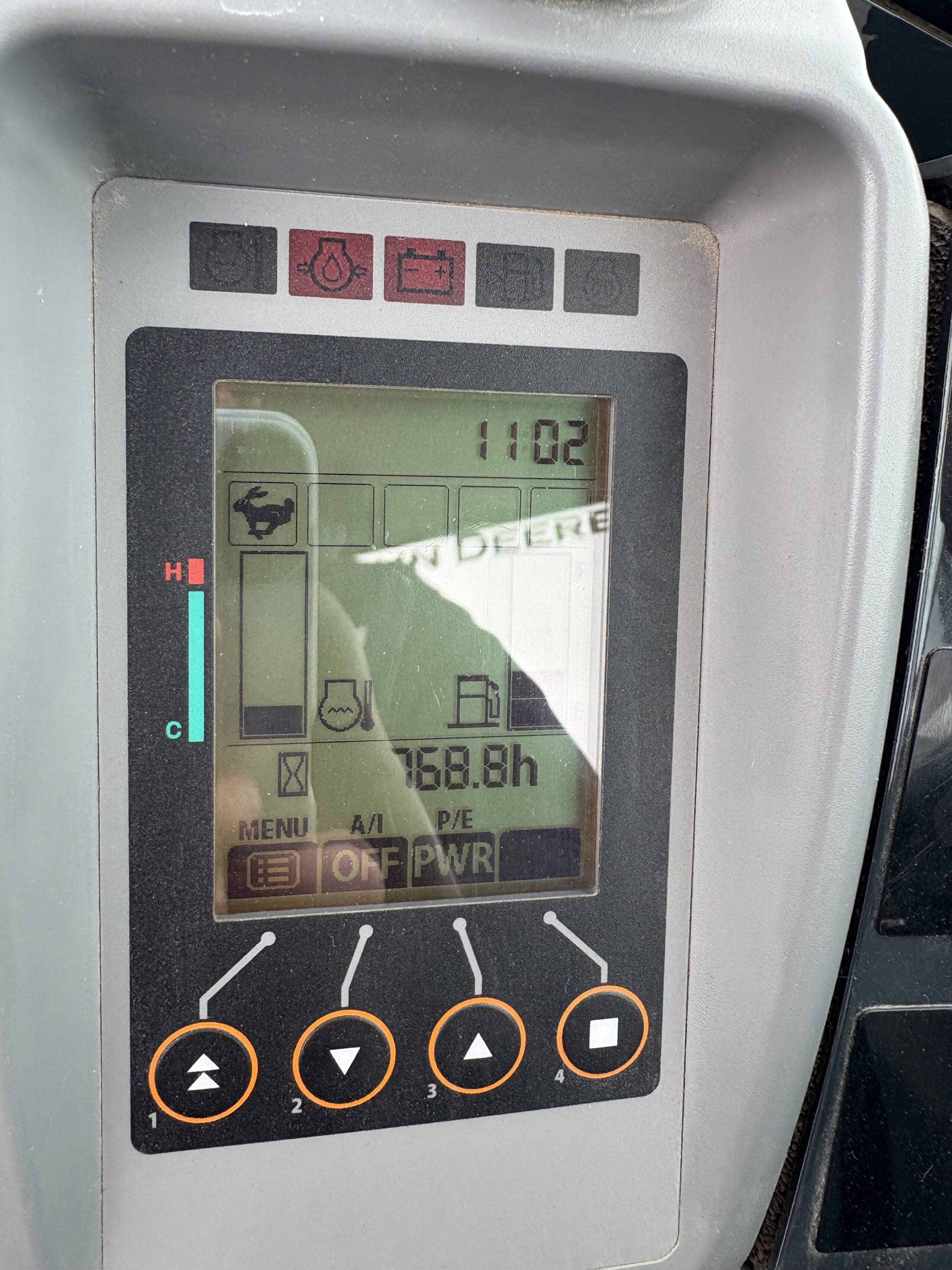 Gros plan d'un écran numérique de tracteur John Deere montrant diverses icônes, un voyant d'avertissement de batterie, une jauge de température, 768,8 heures de fonctionnement, l'heure 11:02 et plusieurs boutons de commande sous l'écran.