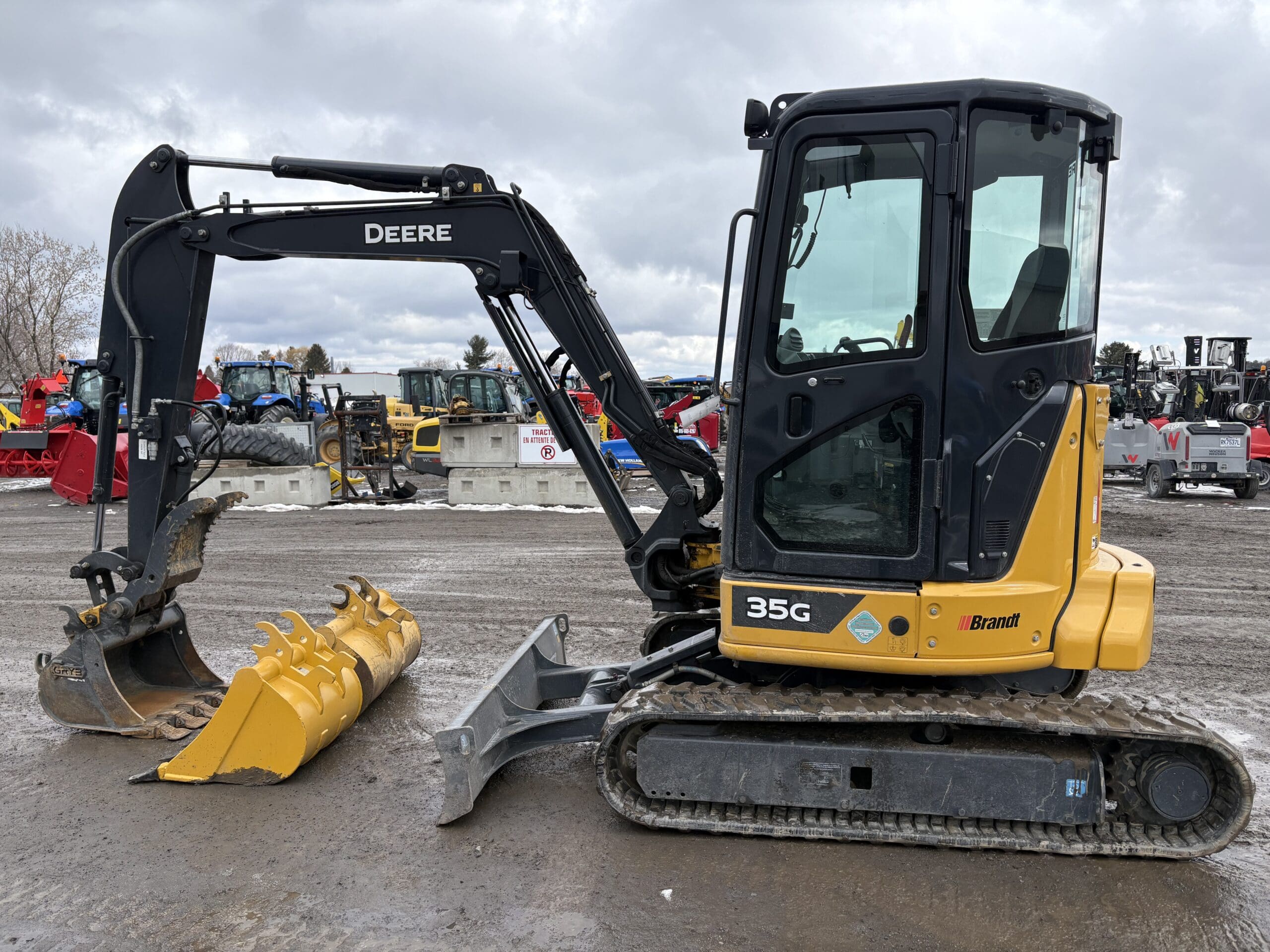 Une excavatrice compacte John Deere 35G jaune et noire avec une chenille métallique et un godet est garée sur un terrain en gravier, avec divers équipements de construction visibles en arrière-plan par une journée nuageuse.