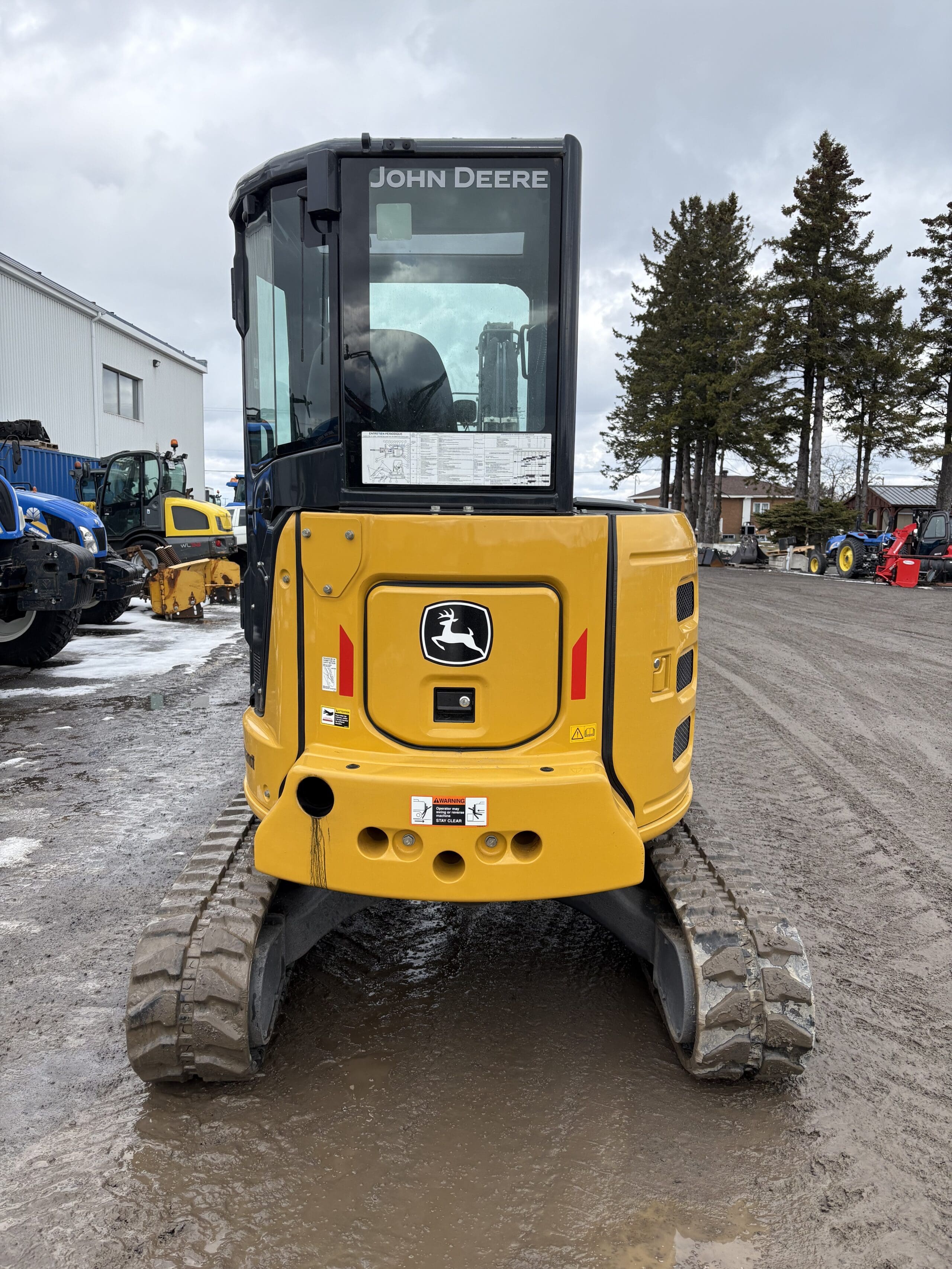 Une pelle compacte John Deere jaune est garée sur un terrain boueux, vue de l'arrière. On distingue des arbres, des bâtiments et d'autres engins de chantier en arrière-plan.