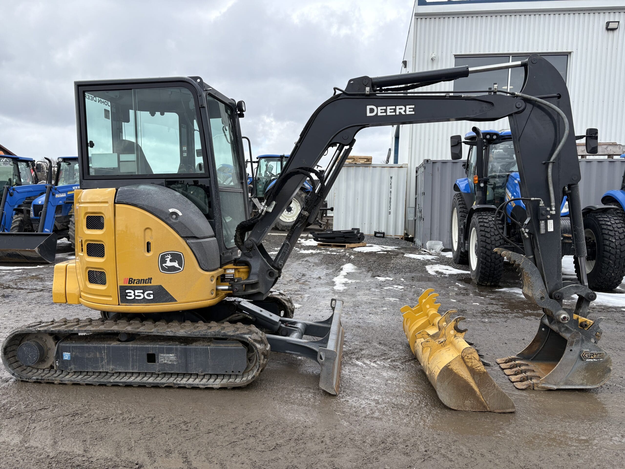 Une excavatrice compacte John Deere 35G jaune et noire avec un godet de creusement est garée sur un sol boueux près de tracteurs bleus et de bâtiments industriels.