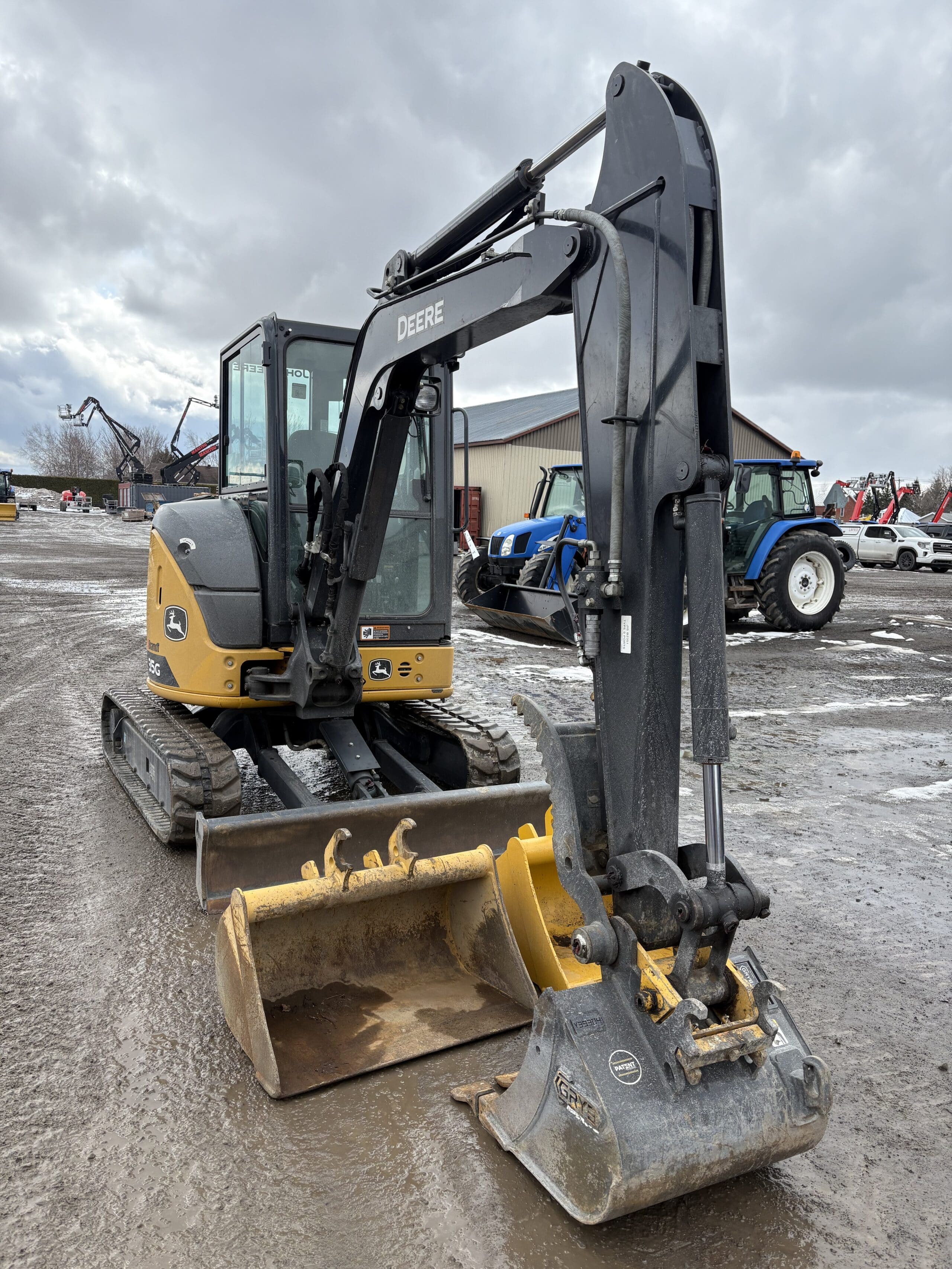 Une excavatrice John Deere jaune et noire avec un grand godet est garée sur un terrain boueux, avec d'autres machines et un tracteur bleu visibles en arrière-plan sous un ciel nuageux.