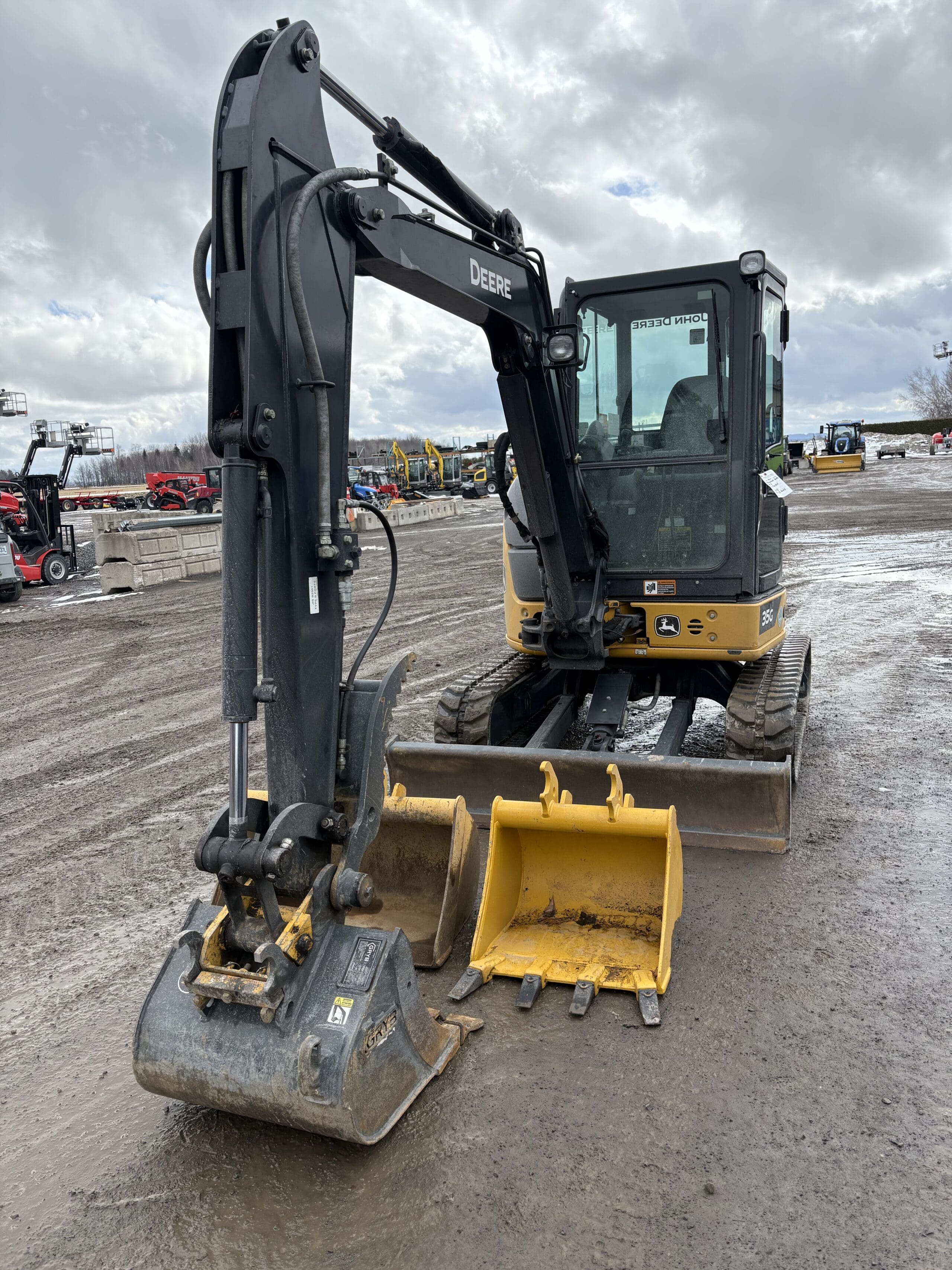 Une mini-pelle John Deere jaune et noire, équipée de deux godets avant, est garée sur un terrain boueux sous un ciel nuageux. D'autres engins de chantier sont visibles en arrière-plan.