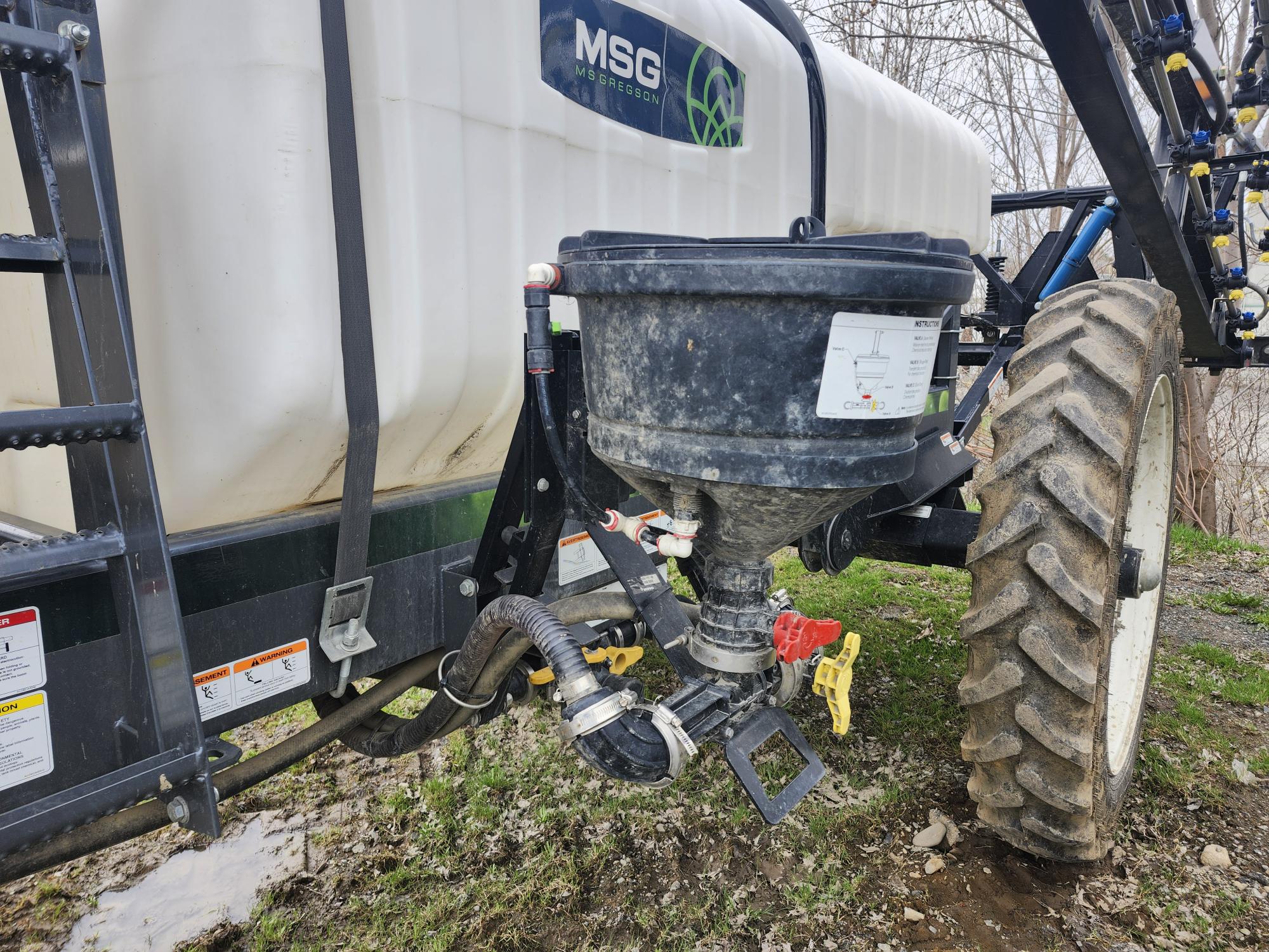 Gros plan d'un équipement agricole attaché à un tracteur, montrant un réservoir chimique blanc, une grande trémie noire, divers tuyaux et vannes et un pneu de tracteur robuste sur un sol herbeux et irrégulier.