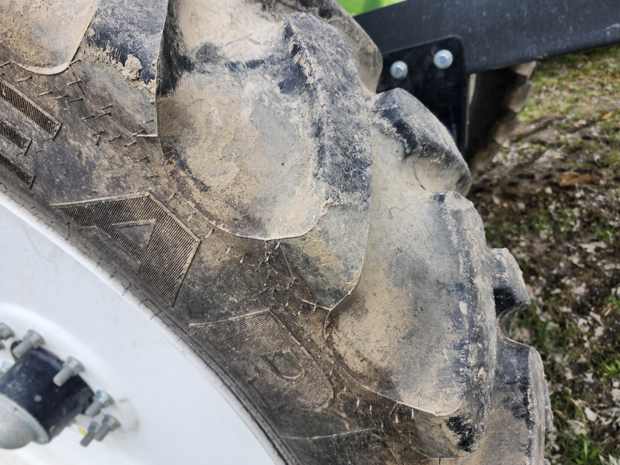 Gros plan d'un pneu de tracteur sale, dont les épaisses bandes de roulement sont couvertes de boue. Le moyeu de roue en métal blanc avec ses boulons est partiellement visible, et l'arrière-plan laisse apparaître de l'herbe et de la terre.