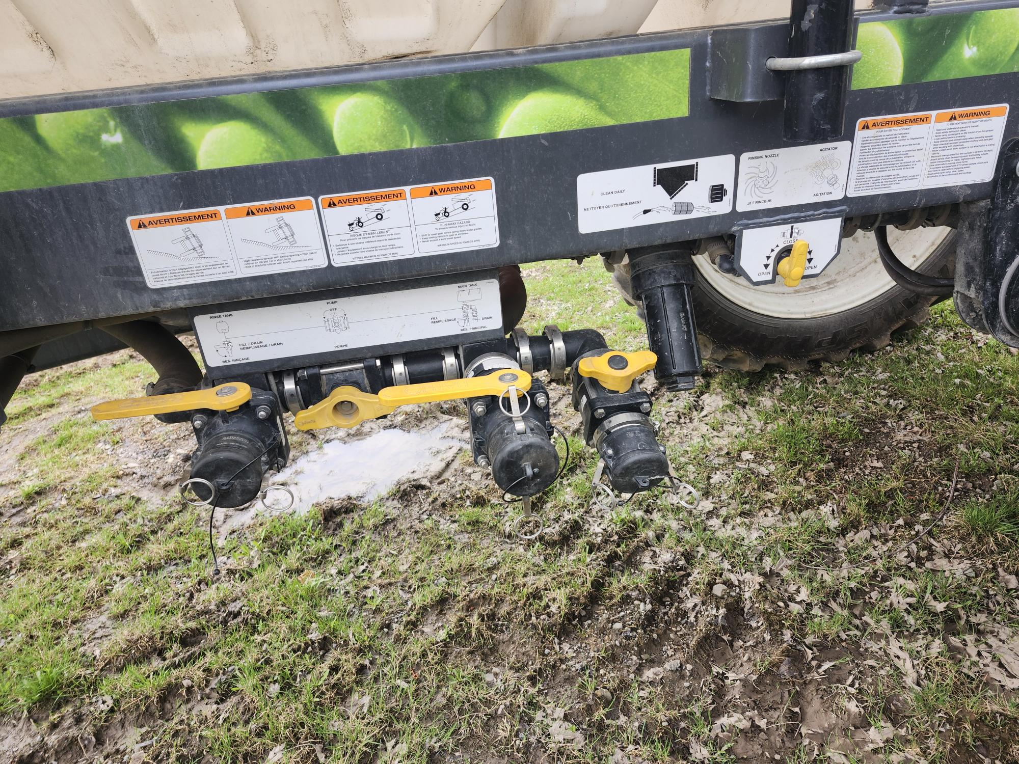 Plusieurs commandes de vannes à poignée jaune et raccords de tuyauterie noirs montés sur du matériel agricole au-dessus de l'herbe boueuse, avec des étiquettes d'instructions et d'avertissement fixées au-dessus des vannes.