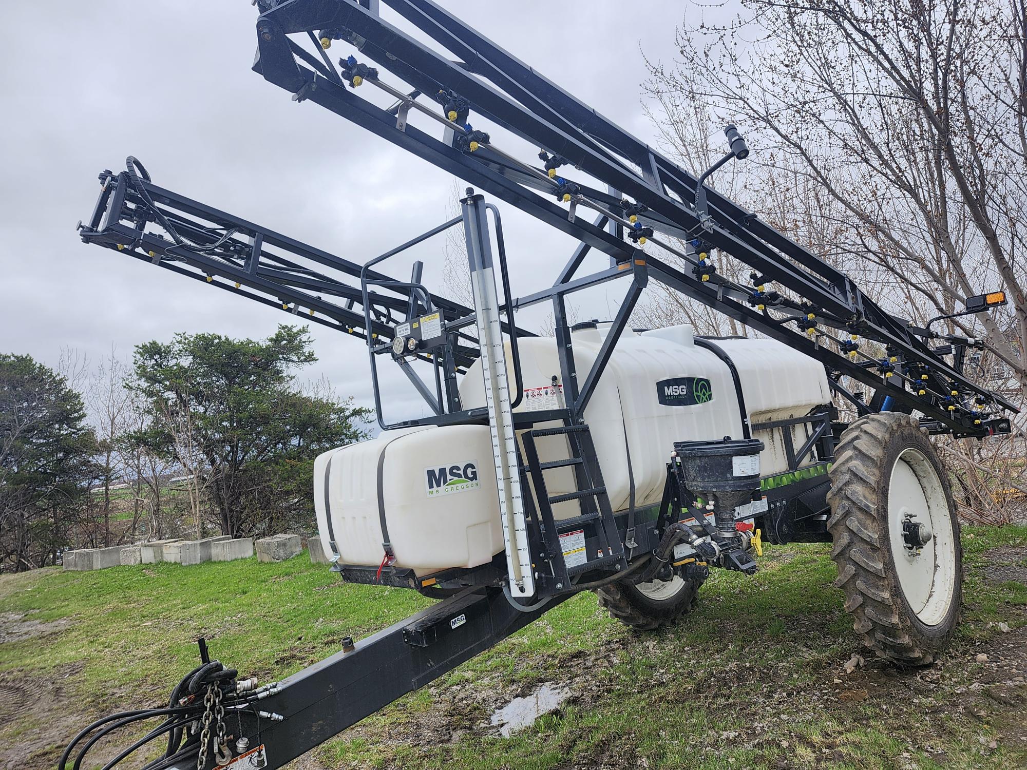 Un grand pulvérisateur agricole équipé de deux rampes allongées est stationné sur l'herbe. La cuve blanche est étiquetée « MSG » et l'engin est équipé de grandes roues. Des arbres et un ciel nuageux apparaissent en arrière-plan.
