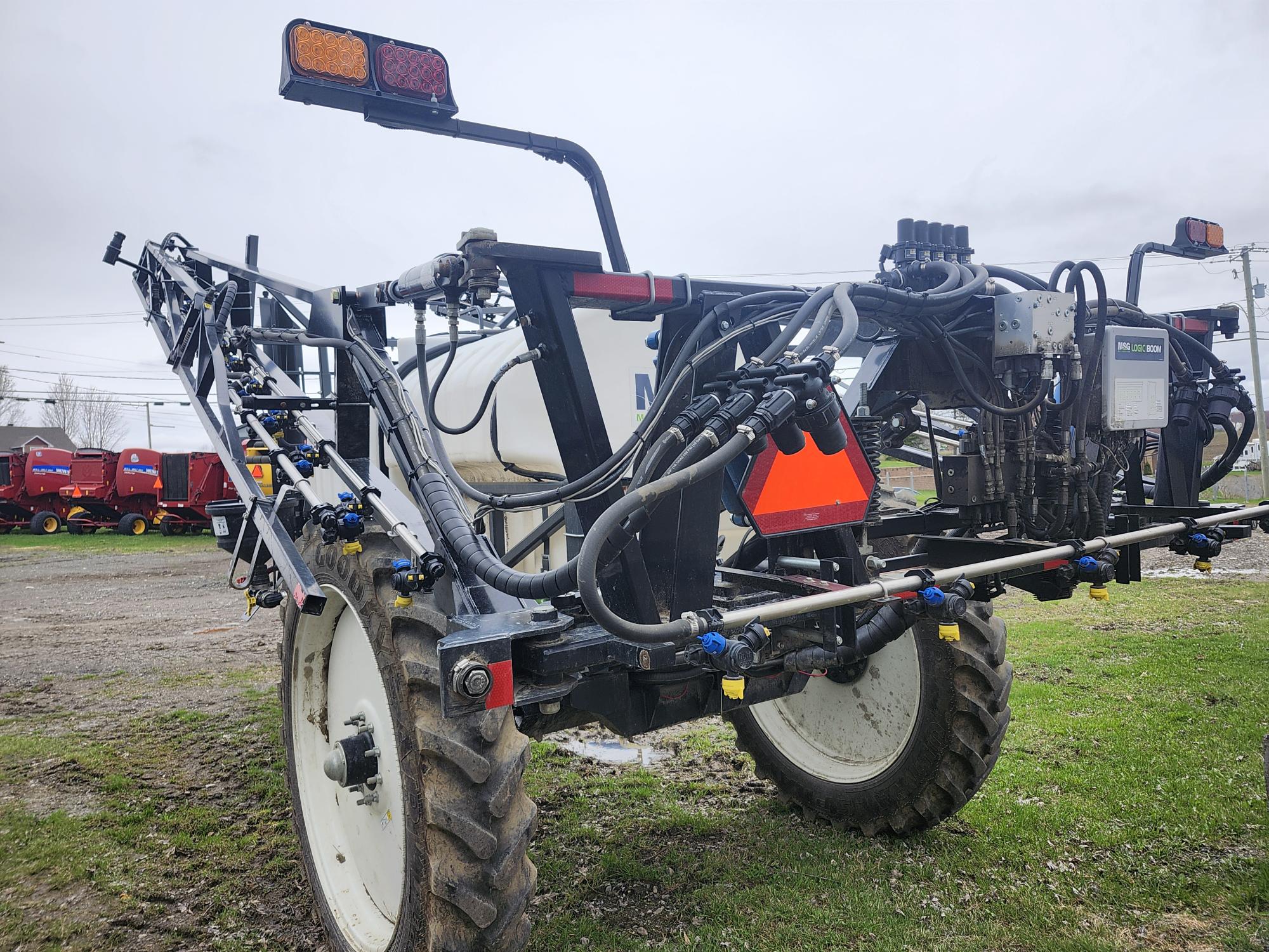 Vue rapprochée d'un pulvérisateur de terrain avec de gros pneus, plusieurs tuyaux, buses et commandes électroniques, garé sur l'herbe à proximité de plusieurs machines agricoles rouges par temps nuageux.