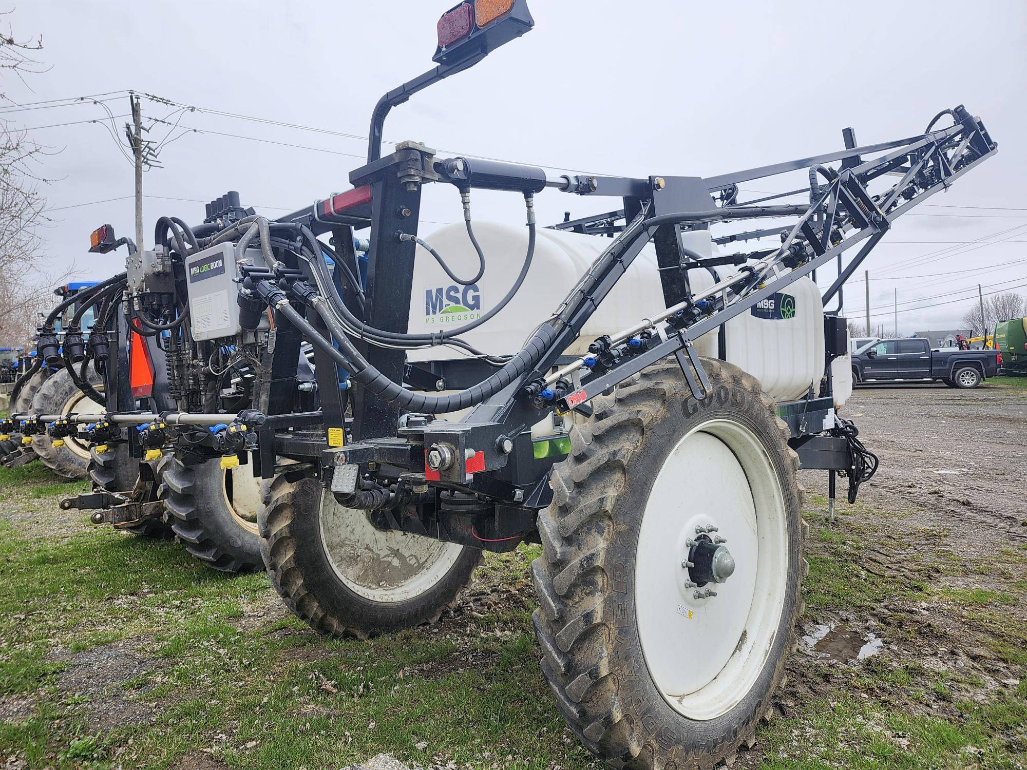 Un grand pulvérisateur agricole, équipé d'une cuve et de rampes, est stationné à l'extérieur sur de l'herbe et de la terre. Le pulvérisateur est doté de gros pneus, du logo MSG, de tuyaux et d'équipements visibles, avec un ciel nuageux et des véhicules en arrière-plan.