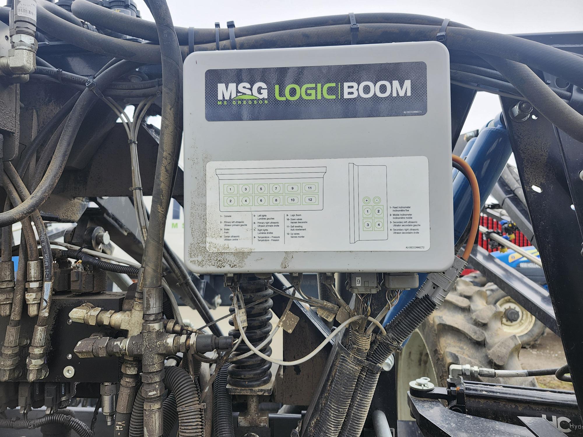 Vue rapprochée d'un boîtier de commande MSG Logic Boom monté sur une machine agricole, entouré de câbles, de fils et de tuyaux hydrauliques, avec une partie d'un pneu de tracteur visible en arrière-plan.